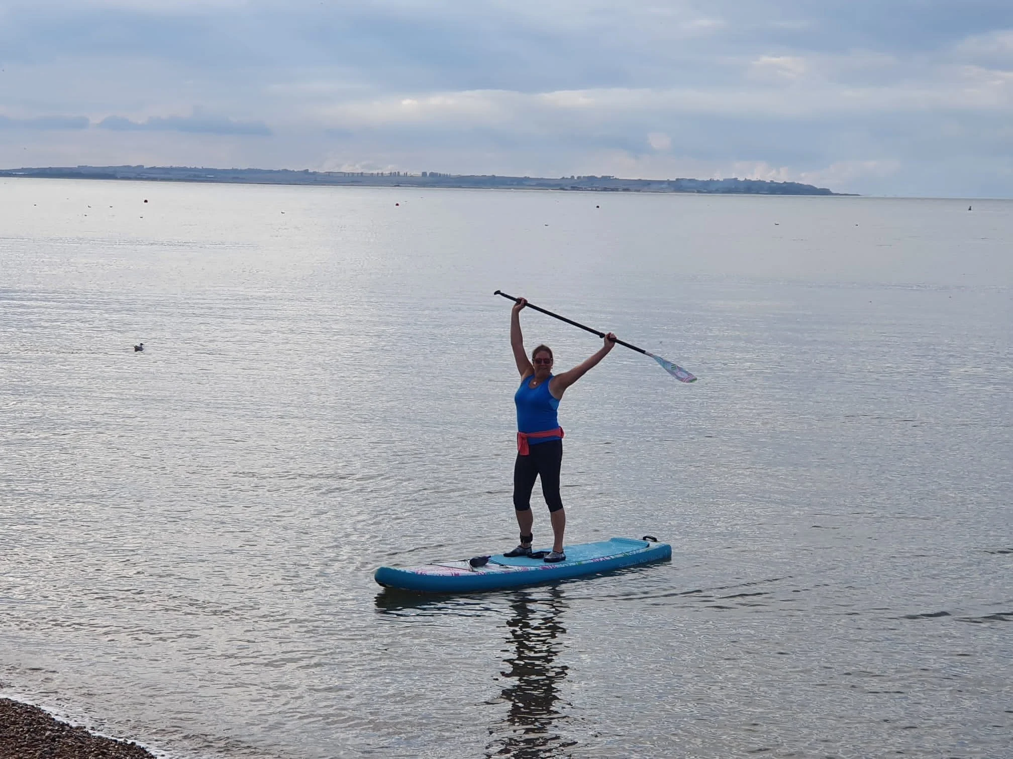 Woman standing on a paddleboard on calm water, holding a paddle overhead with both arms raised.