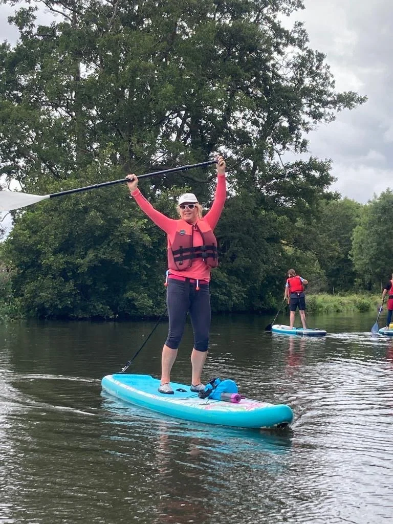 Embodiment coach standing on a paddleboard on a peaceful river with arms raised in celebration, wearing a life vest and smiling.