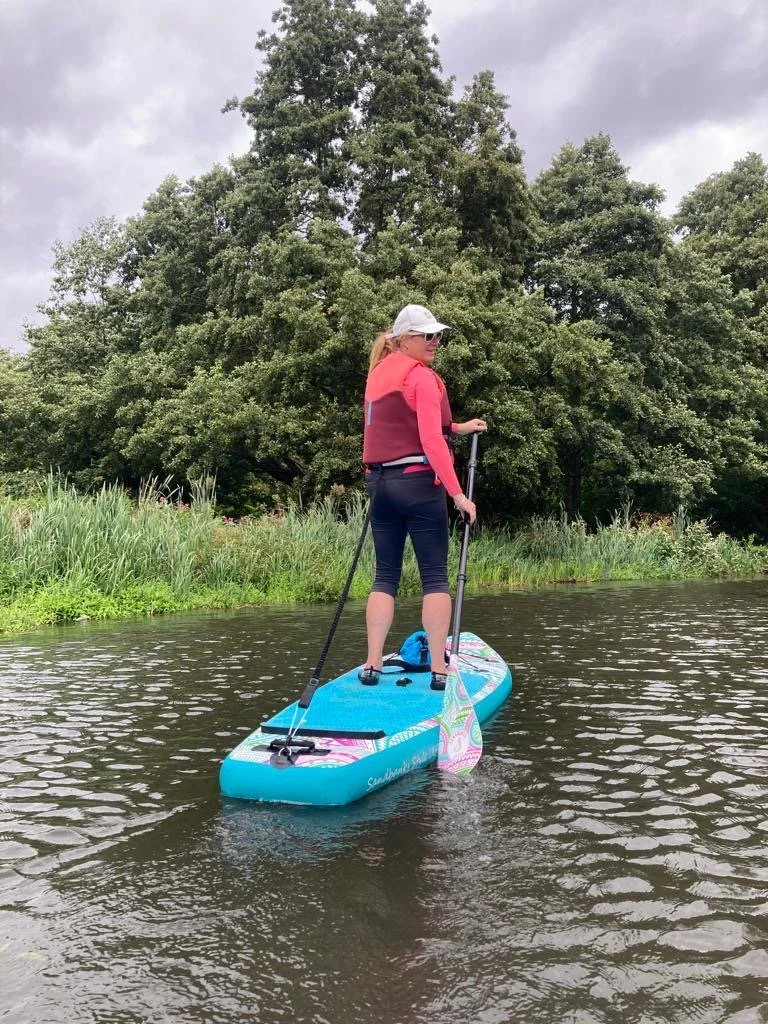 Woman standing on a paddleboard on a calm river with her arms raised holding a paddle, wearing a life vest and smiling, surrounded by trees.