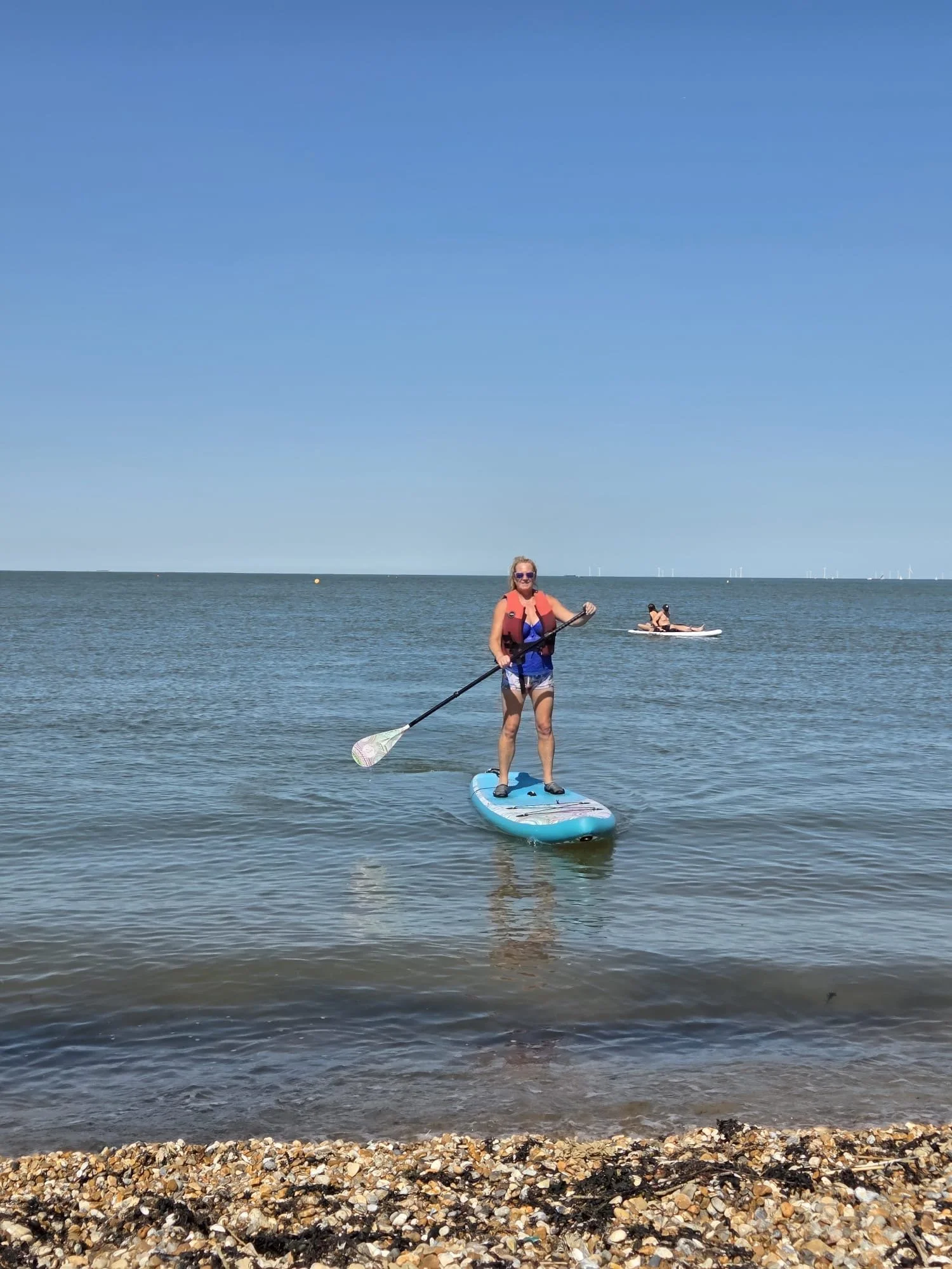 A woman in a red life vest paddleboards on calm ocean water under a clear blue sky. Another paddleboarder is visible in the distance. The scene is peaceful.