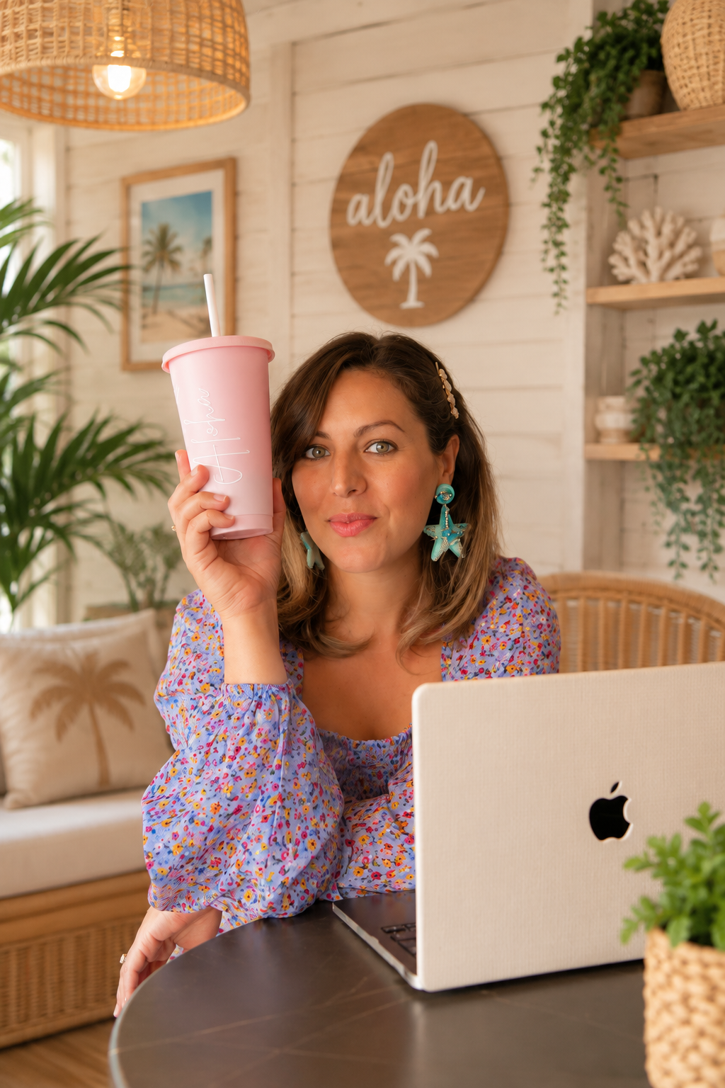 A woman with shoulder-length brown hair and starfish earrings sitting at a table with a laptop, holding a pink cup with a straw, in a tropical-themed, decorated room with a sign that says 'aloha' and palm tree artwork.