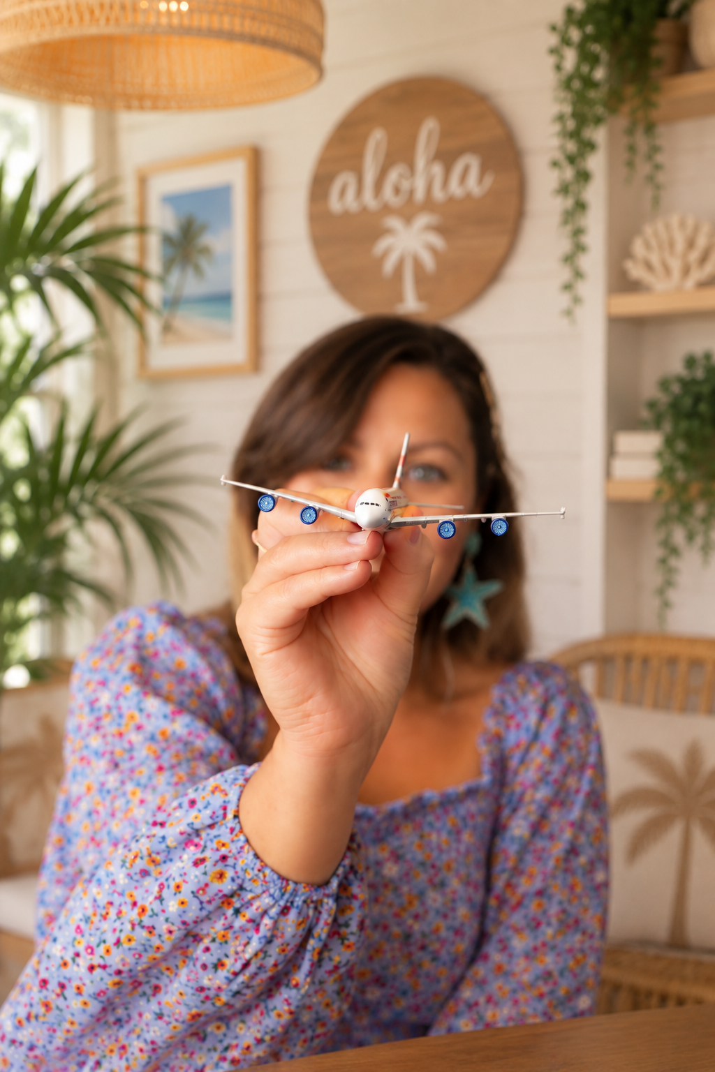 A woman holding a toy airplane in front of her face with a tropical-themed decor background, including a sign that says 'aloha' with a palm tree, plants, and beach-themed artwork.
