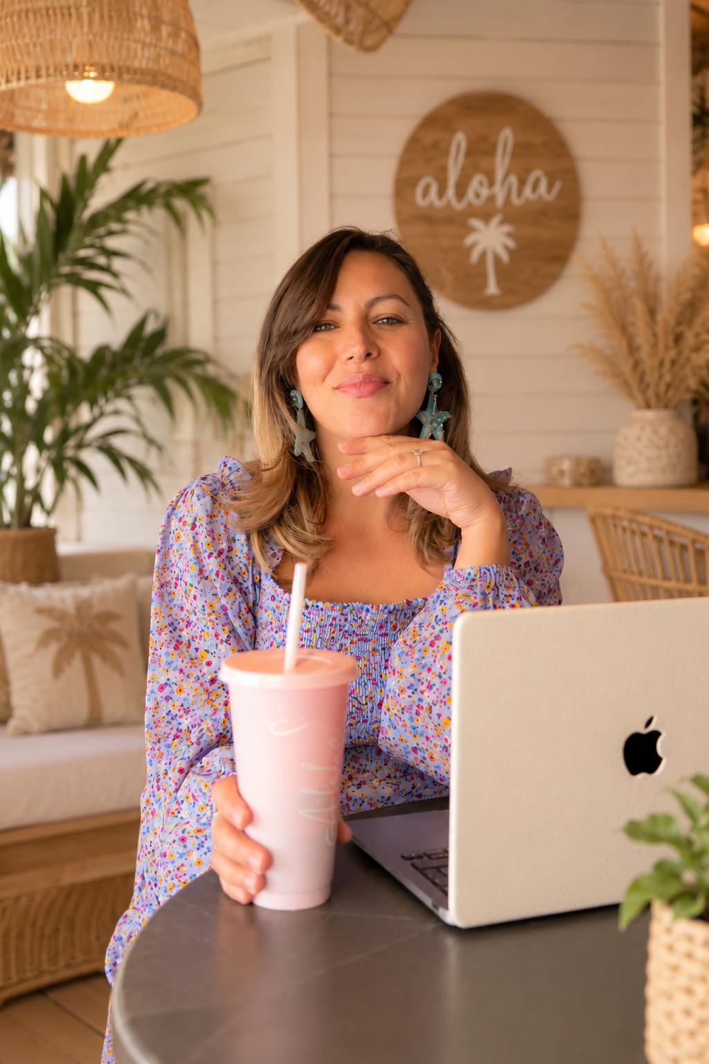 A social media marketer sitting at a table with a laptop, holding a pink drink with a straw, in a cozy, tropical-themed cafe or home interior with plants and a sign that says 'aloha' in the background.