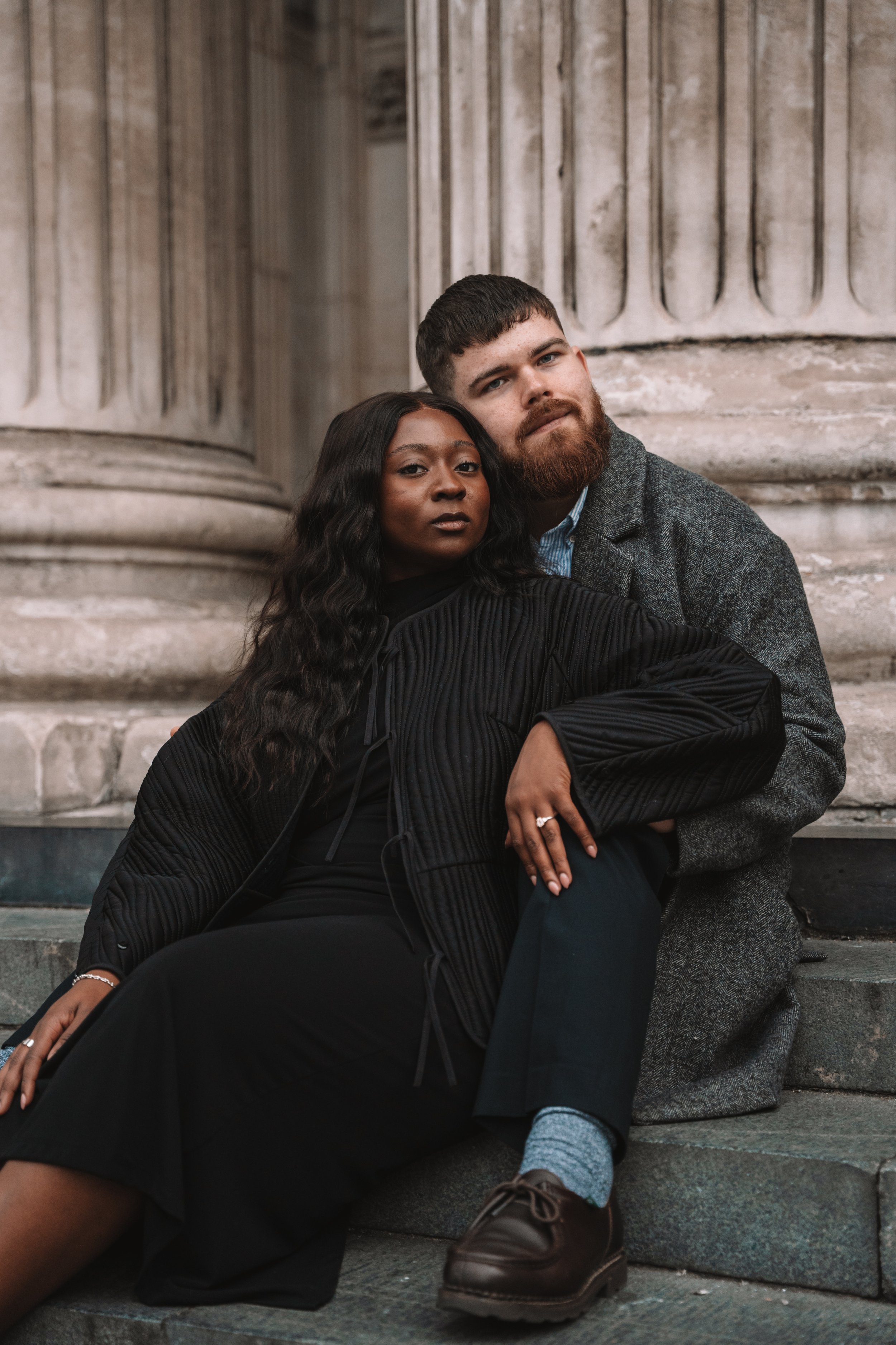 A black woman and a white man sitting on stone steps in front of a classical building with large columns, looking at the camera.