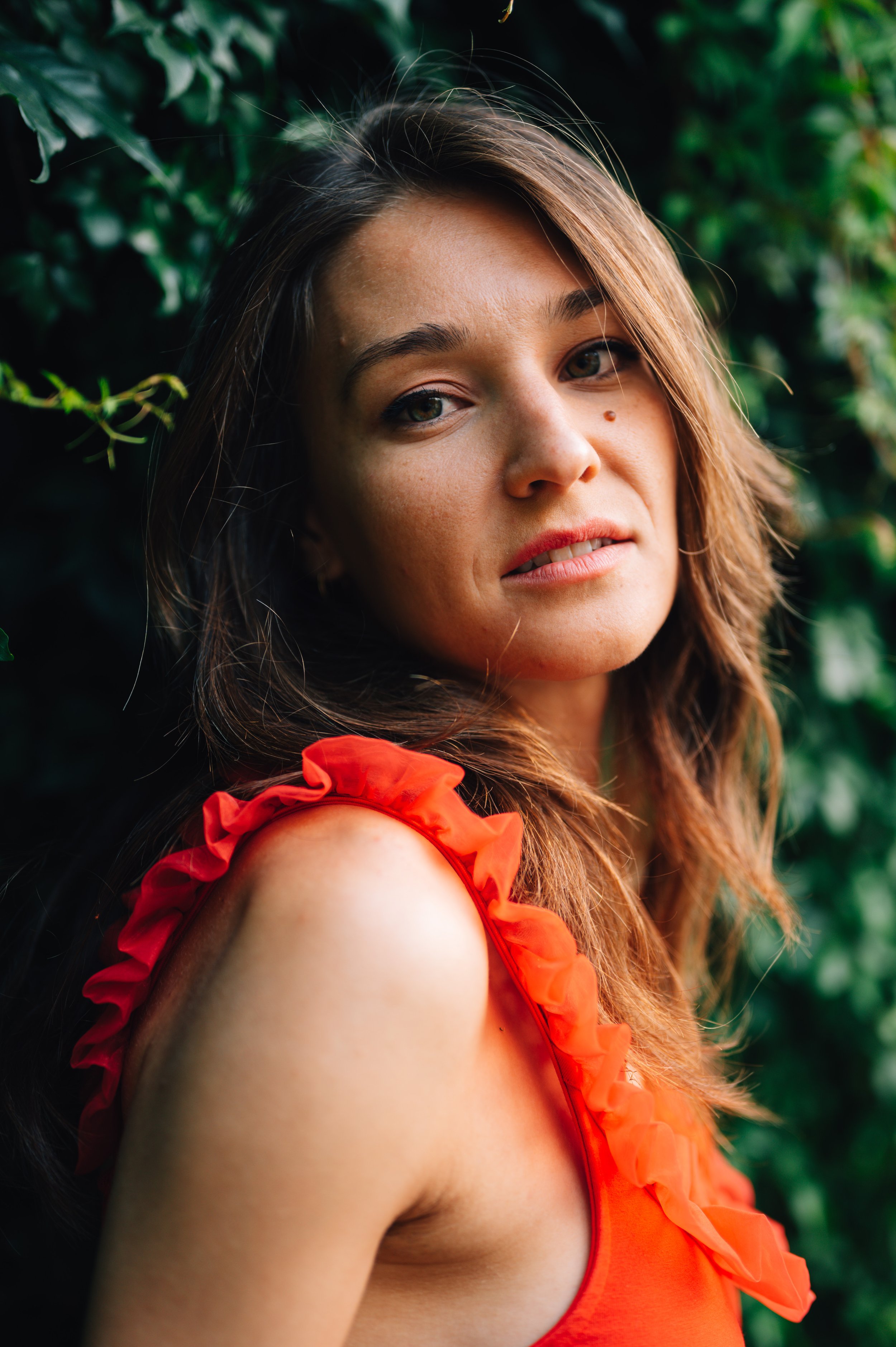 A woman with shoulder-length brown hair and light skin wearing an orange ruffled top, standing outdoors with green foliage in the background.