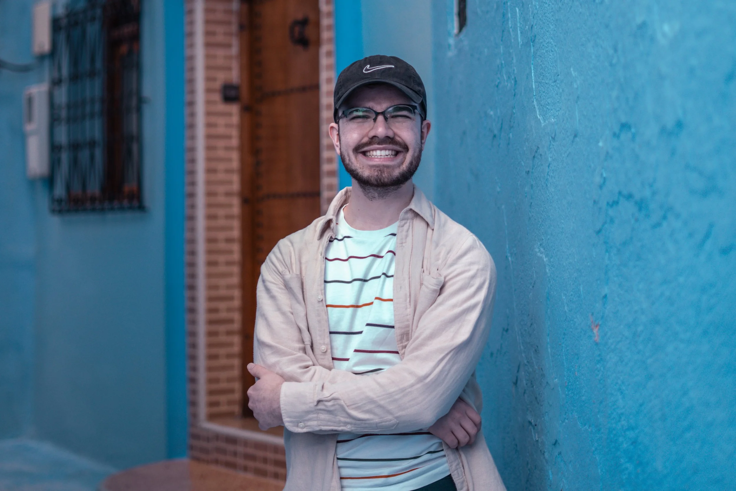 A smiling man with glasses, a black cap, and a beard, wearing a striped shirt and beige jacket, standing against a vibrant blue wall with a wooden door and window in the background.