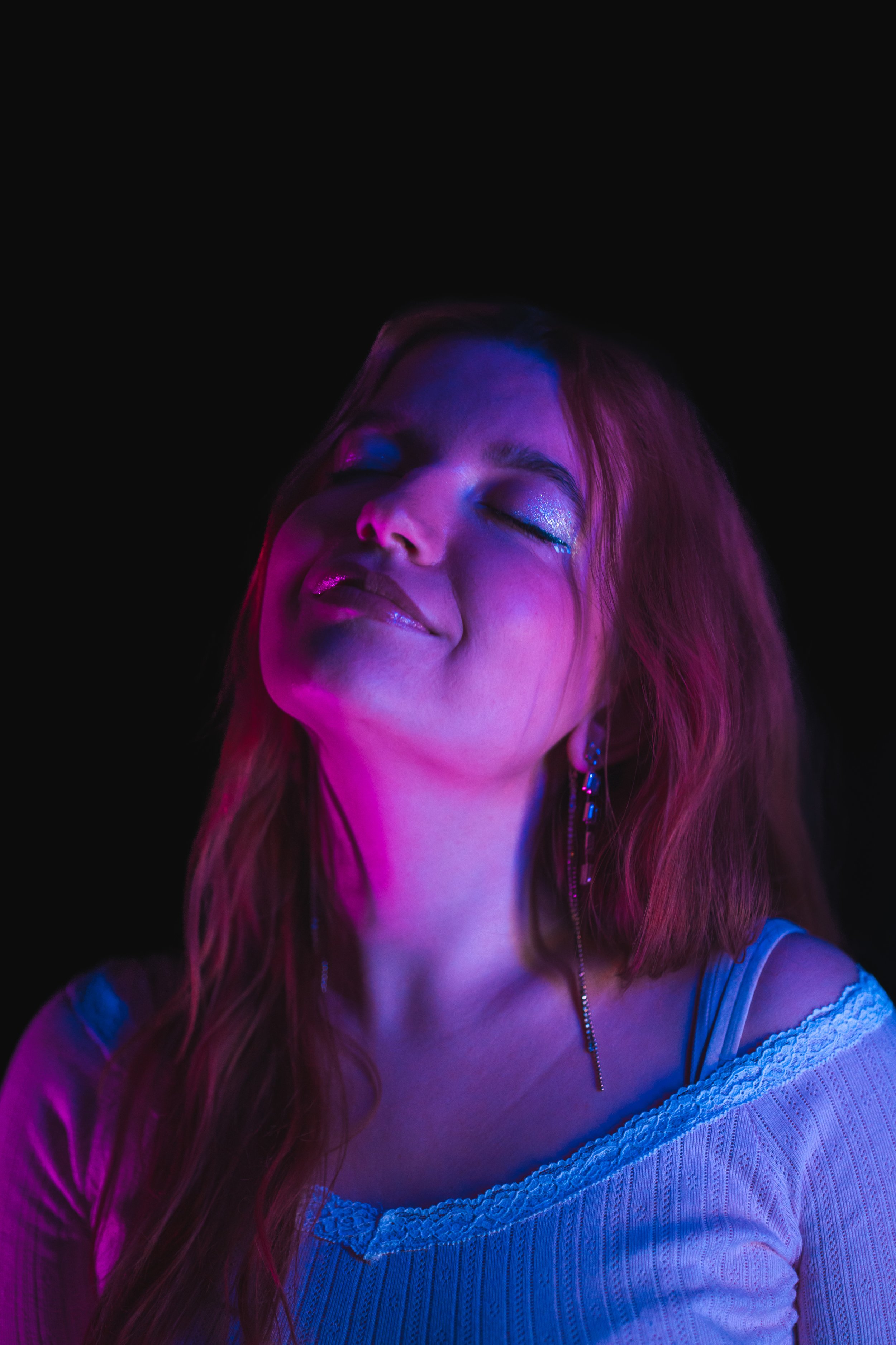 A young woman with closed eyes and a peaceful smile, illuminated with purple and blue neon lights, wearing earrings and a light-colored top with lace detail.