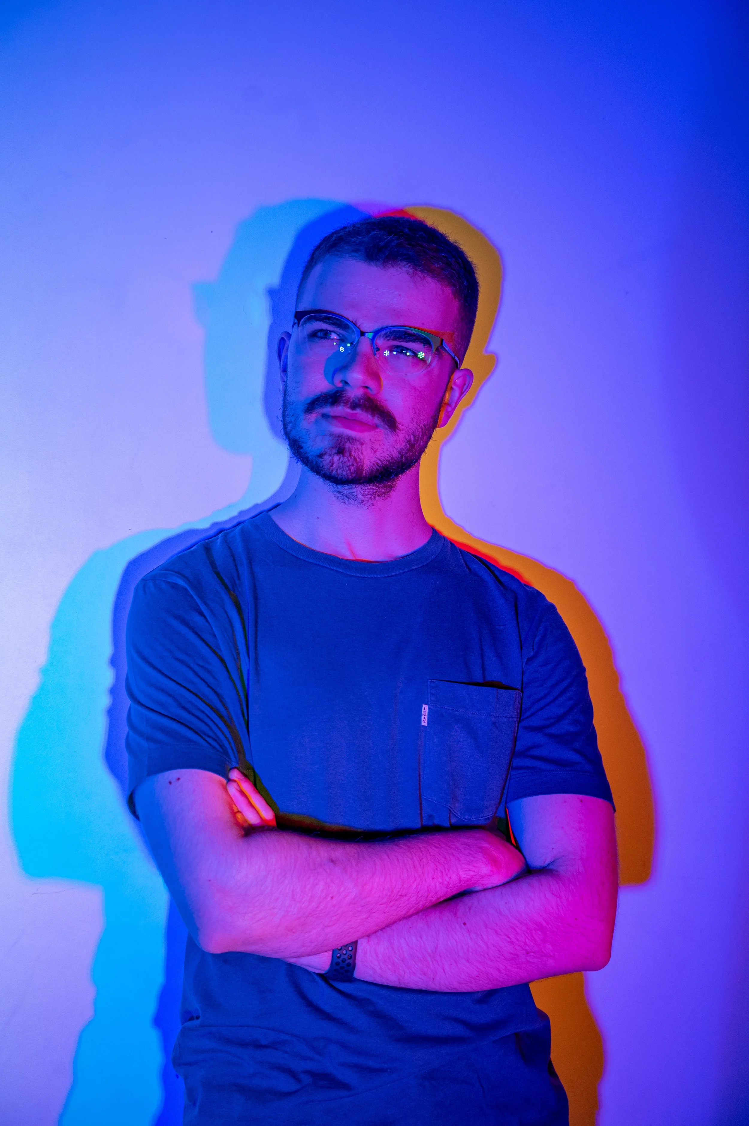 A man with glasses and a beard standing with arms crossed against a wall lit with multicolored lights, creating shadows on the wall.