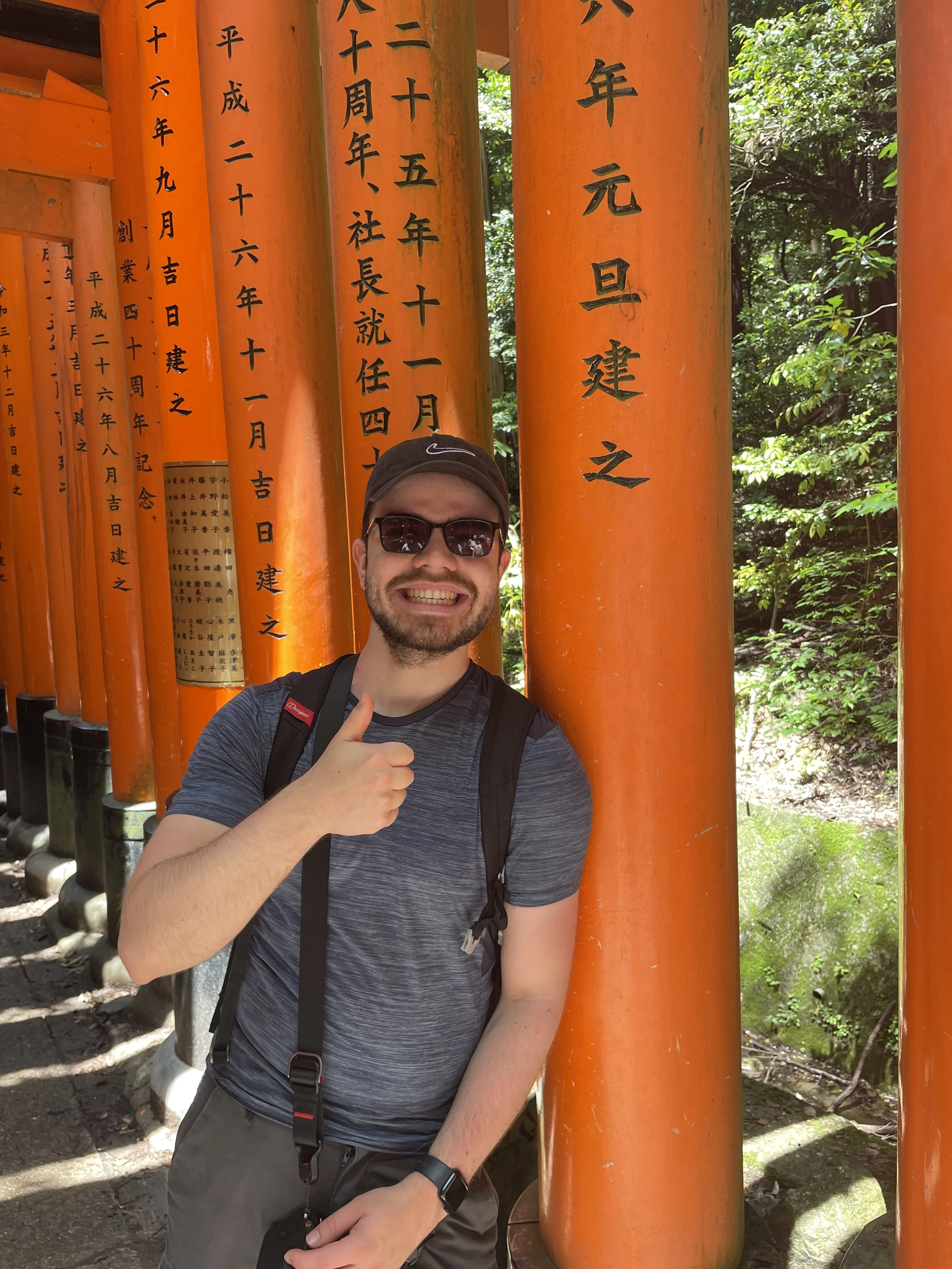 A man wearing sunglasses, a black Nike cap, a gray t-shirt, and a backpack is standing among orange Torii gates with Japanese inscriptions, giving a thumbs-up gesture and smiling at the camera.