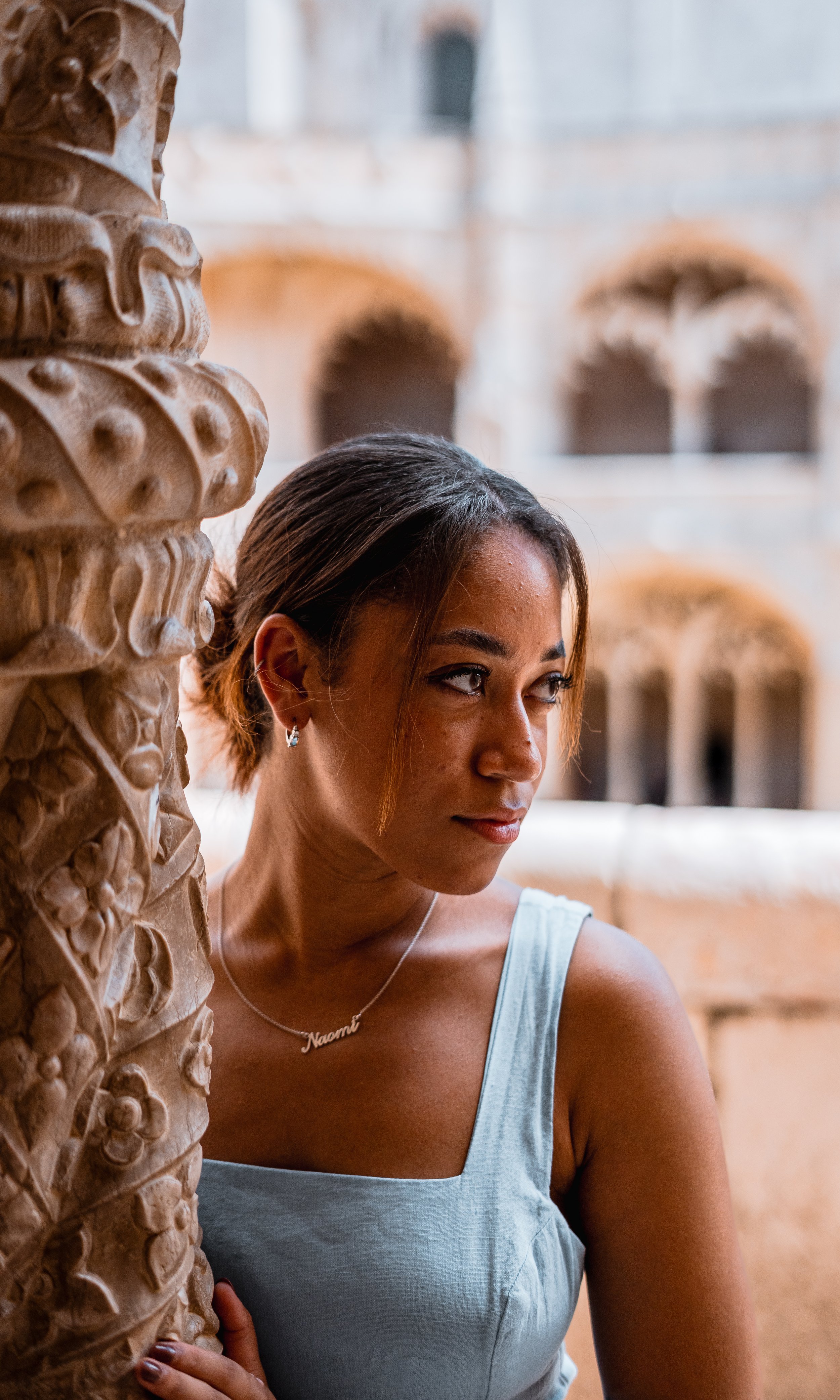 A woman with short dark hair and hoop earrings is standing next to a carved stone column, with architectural arches in the background.