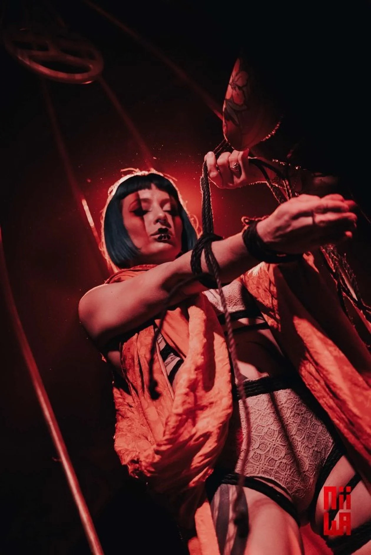 A woman with short black hair and punk style makeup poses with ropes and a fan, illuminated by moody red lighting.