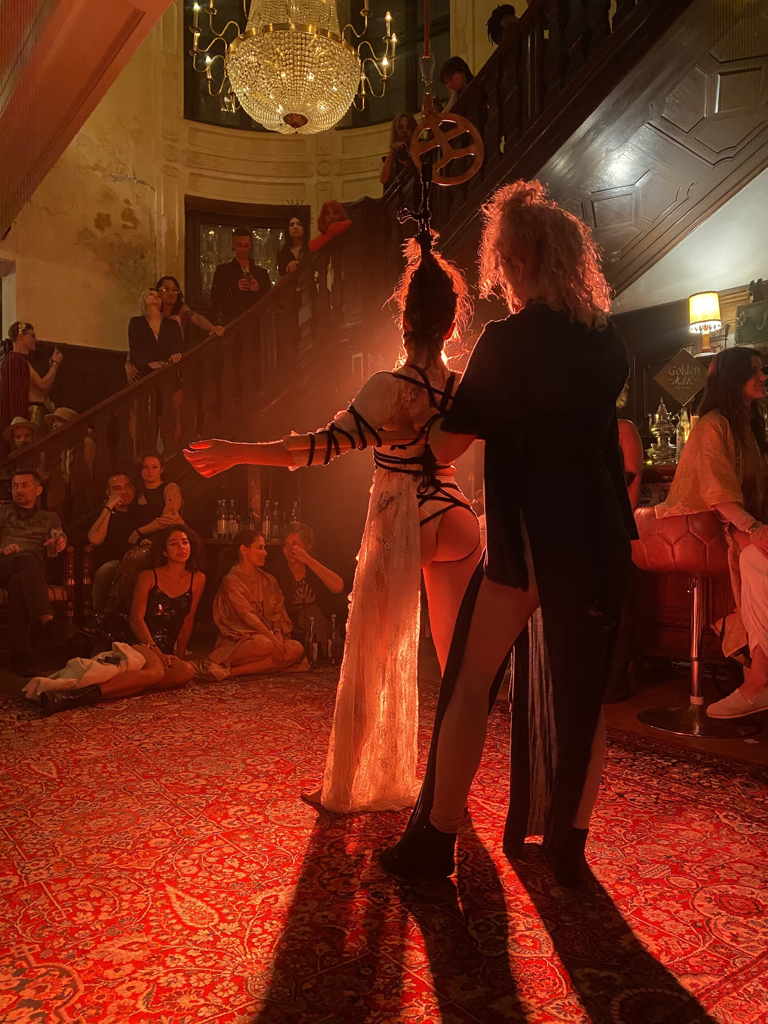 A performer in a revealing costume with black straps and lace, performing in a dimly lit venue with a red carpet. An audience sits on the floor and stands on a staircase, watching the performance.
