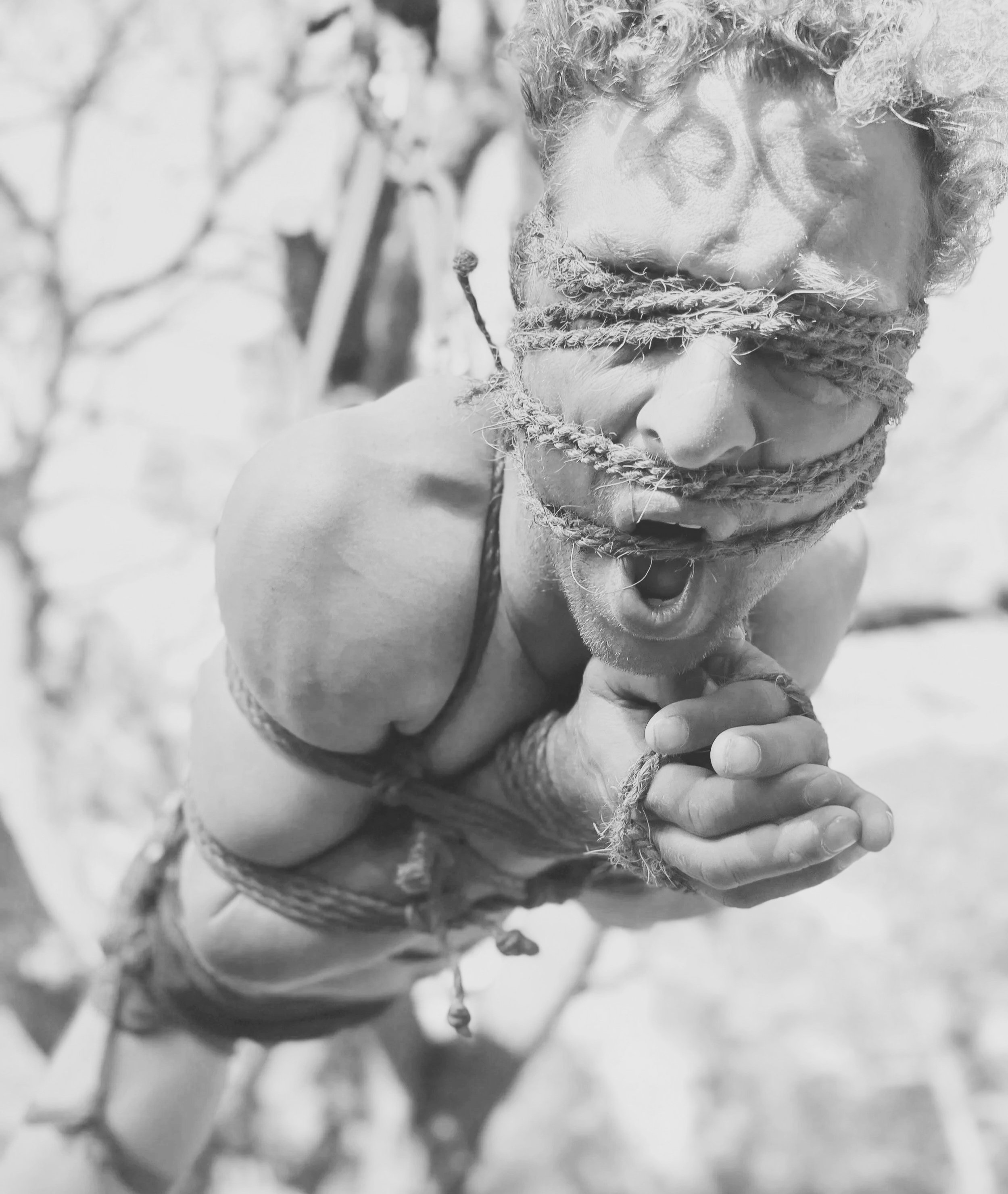 A male person with curly hair is blindfolded with braided rope and is gagged with ropes, with arms tied behind their back, appearing to be in distress or agony outdoors in a black-and-white photo.