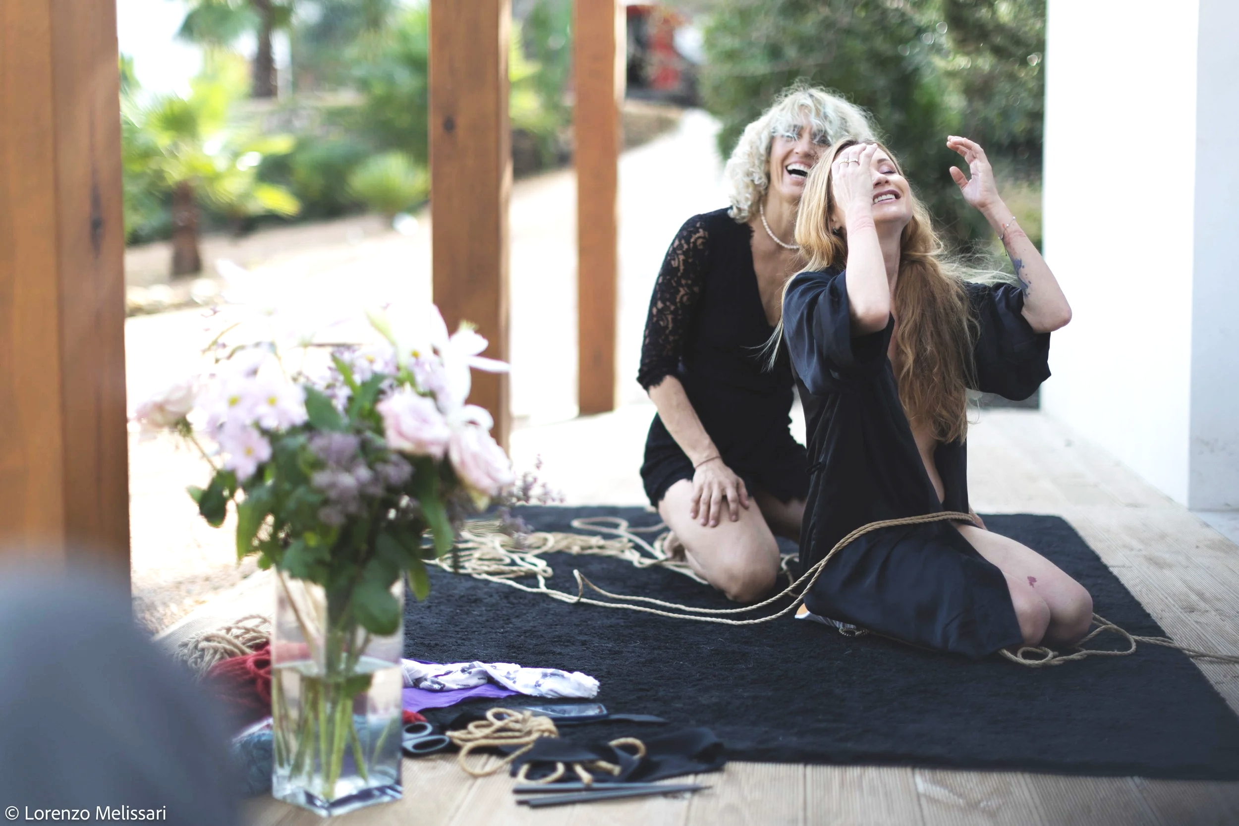 An older woman and a younger woman laughing in an outdoor setting, with the younger woman tied to a rope and sitting on her knees on a black rug, surrounded by flowers and craft supplies.