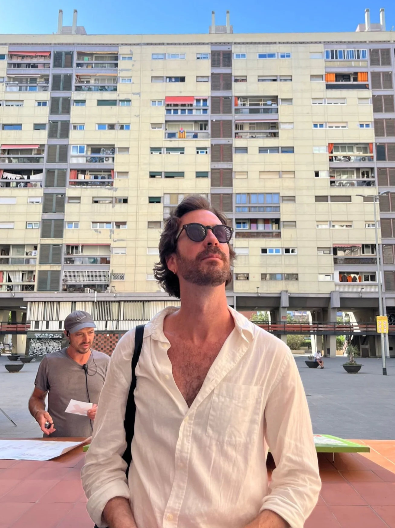 A man with dark, wavy hair and sunglasses stands outdoors in front of a large residential building. A man wearing a gray shirt and cap is in the background, holding papers. The scene appears to be in an urban area with other people and graffiti visible.