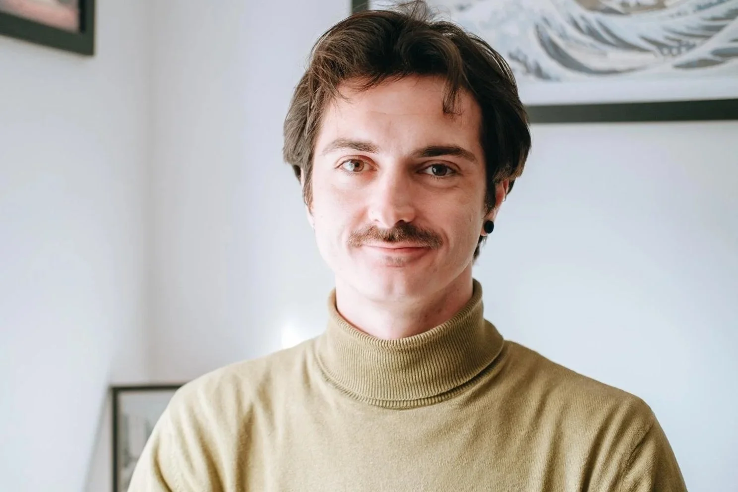 A man with dark hair, a mustache, wearing a khaki turtleneck sweater, standing indoors in front of a white wall with framed artwork.