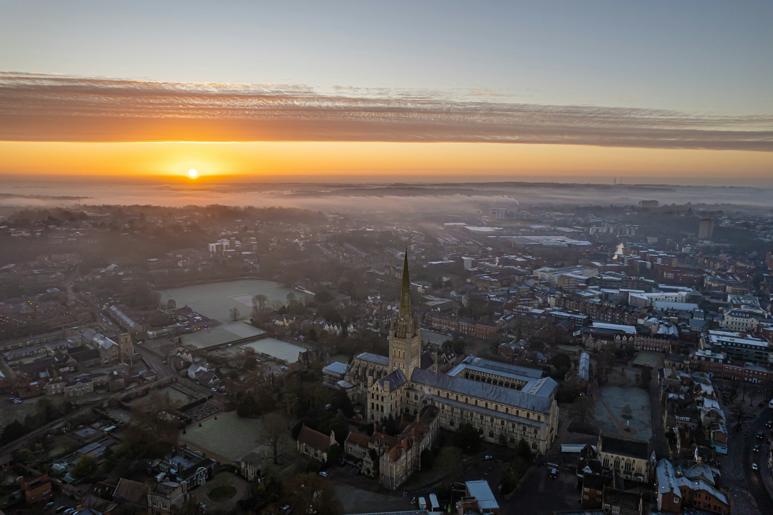 An aerial view of Norwich city at sunrise with the historic cathedral featuring a tall spire at the center, surrounded by residential and commercial buildings, with mist over some areas in the distance.