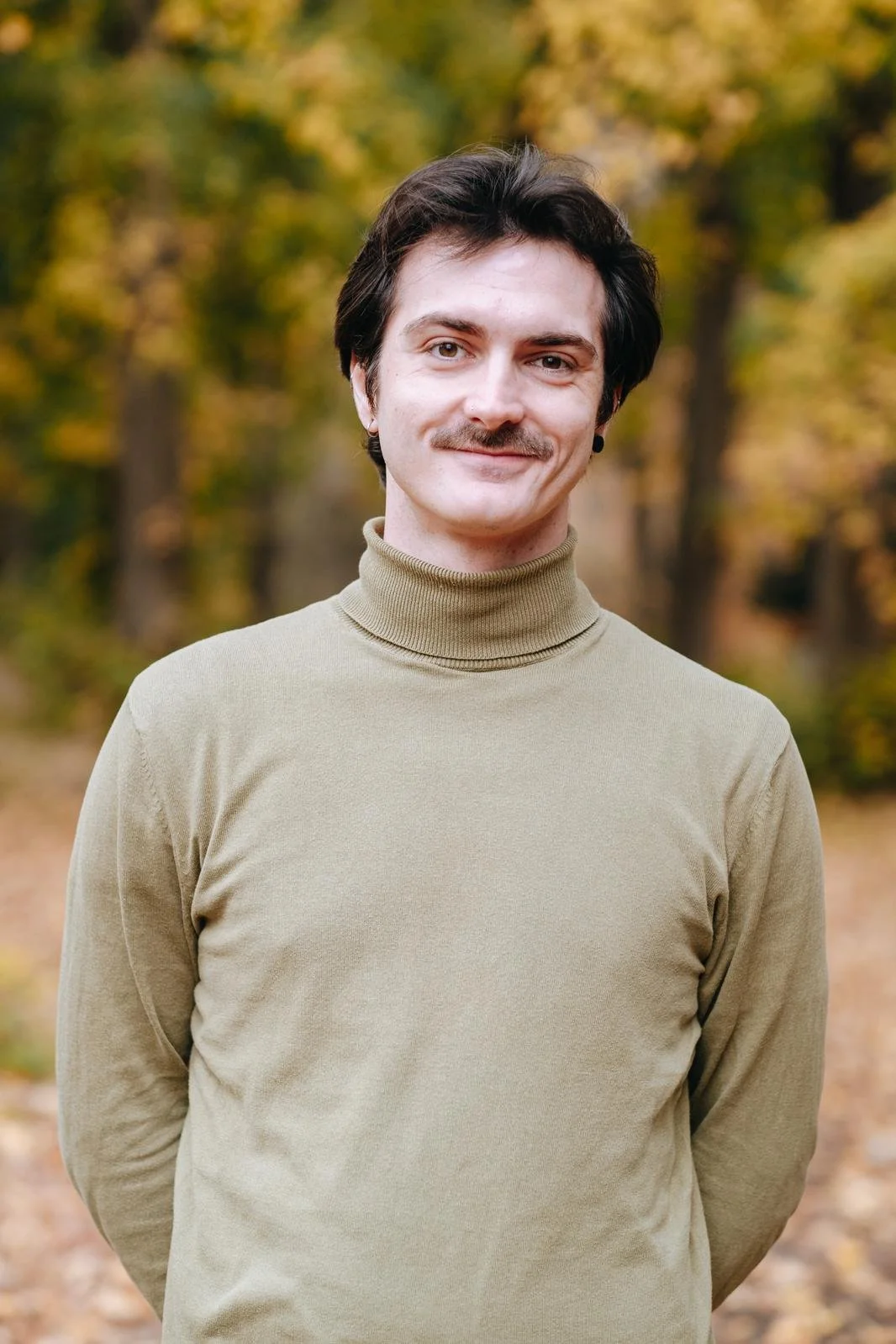 A man standing outdoors in fall, wearing a khaki turtleneck sweater, with a blurred autumn forest background.