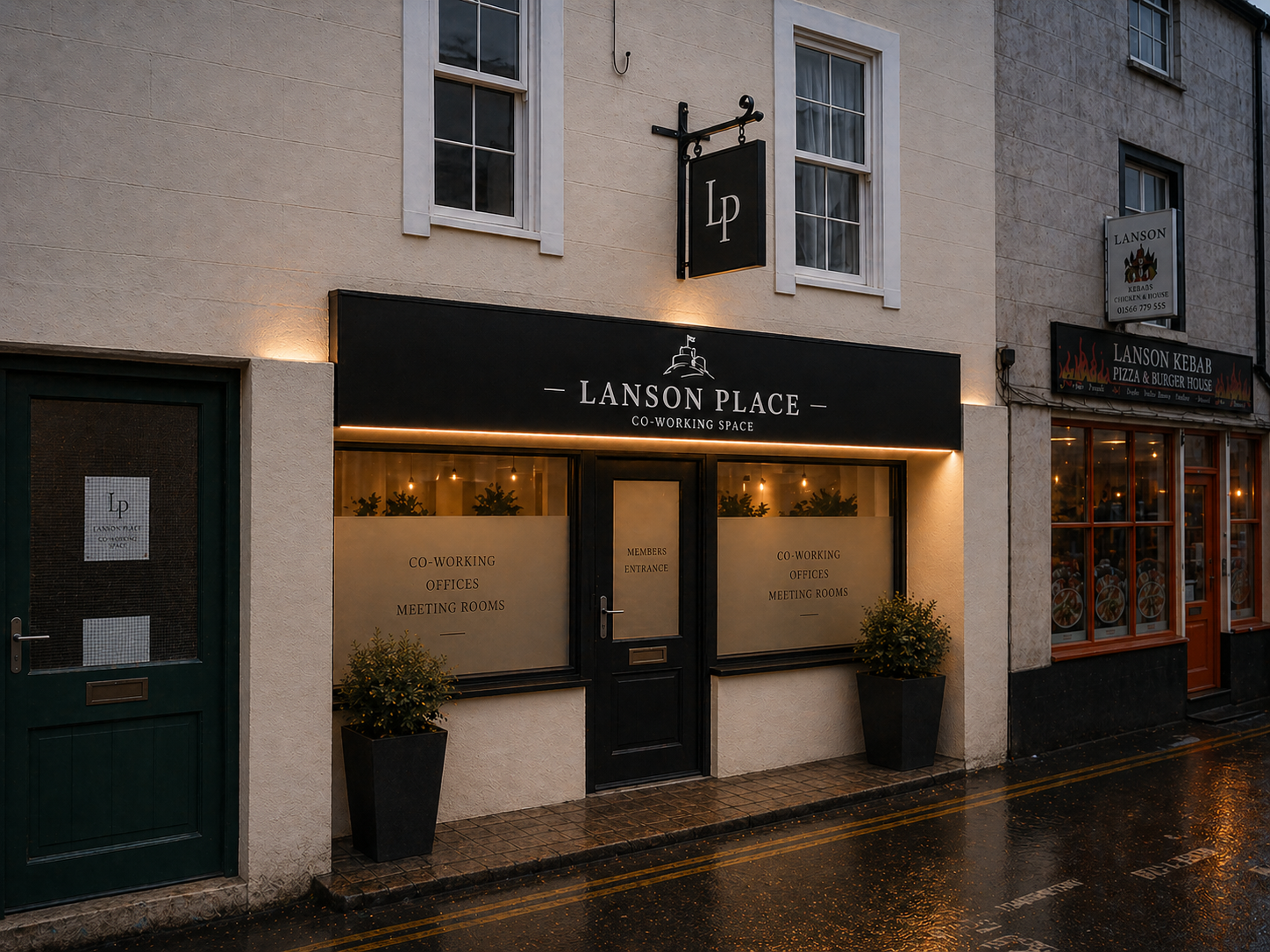 Facade of Lanson Place co-working space with black signboard, glowing warm lights, large windows, potted plants outside, and adjacent shops on a rainy evening.