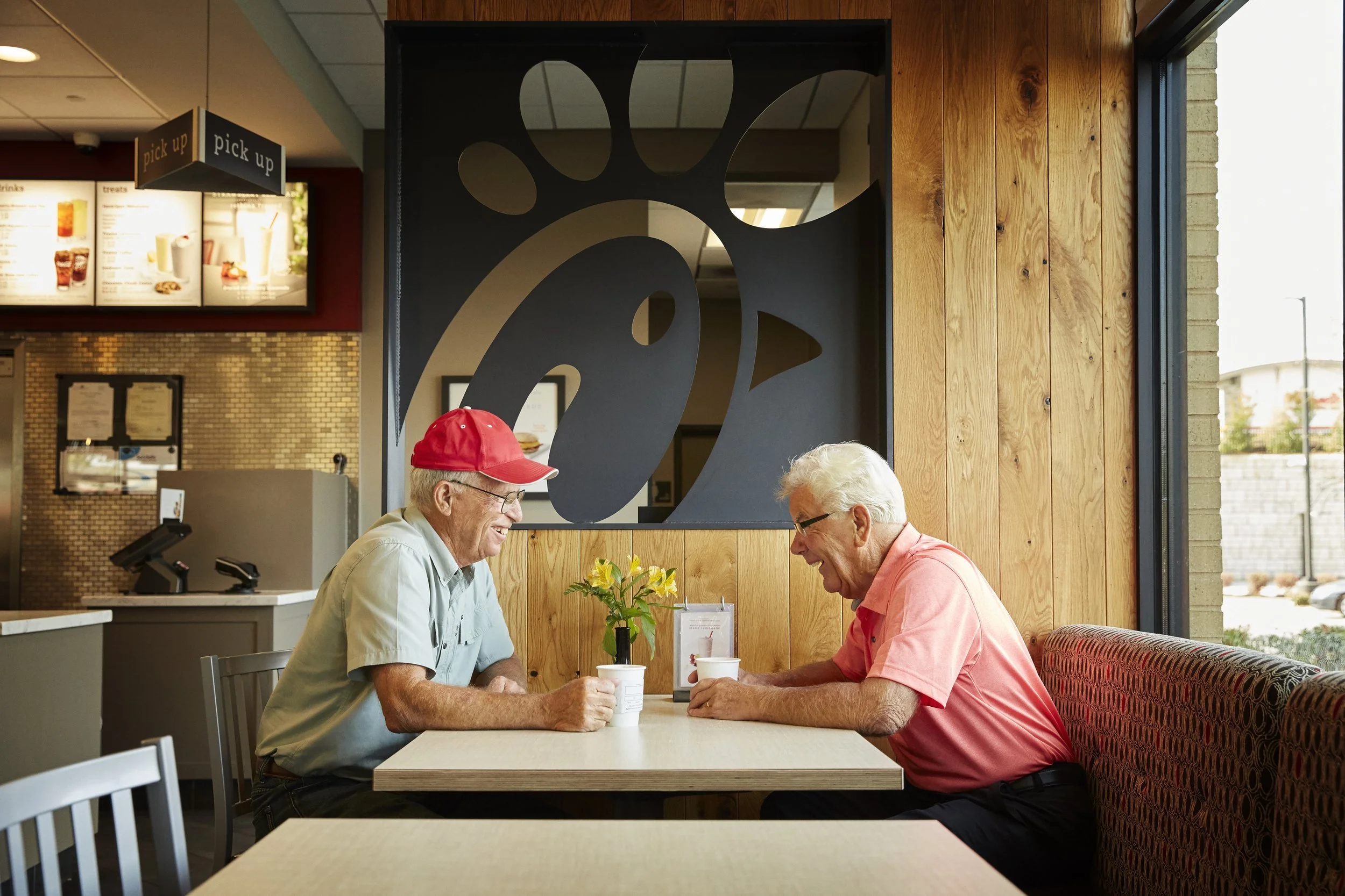 Two elderly men sitting at a table in a restaurant, smiling and talking. One is wearing a red cap and glasses, the other has gray hair. There is a small yellow flower arrangement on the table, and a large Chick-fil-A logo is on the wall behind them.