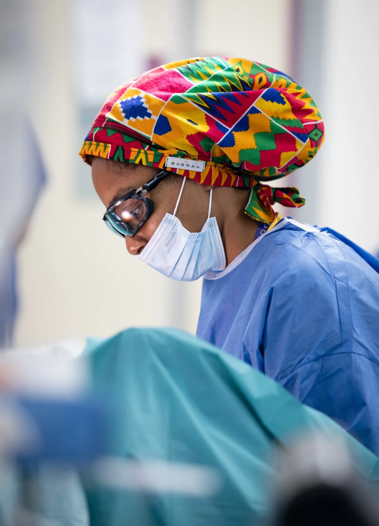 A healthcare professional wearing a colorful headscarf, protective glasses, a mask, and blue medical scrubs working in a hospital setting.