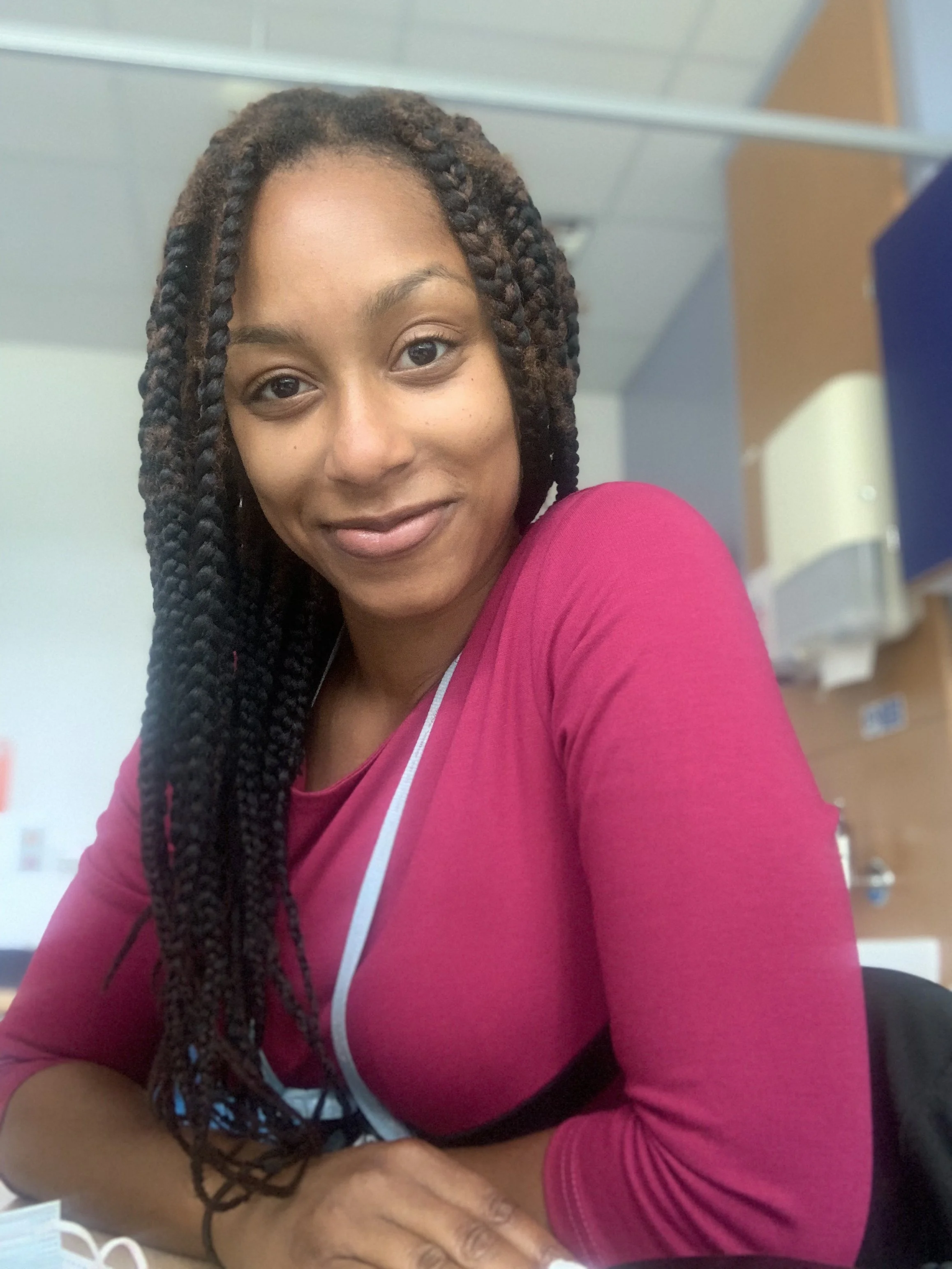 A woman with braided hair smiling at the camera, wearing a pink top, in an indoor setting that appears to be a hospital or medical office.