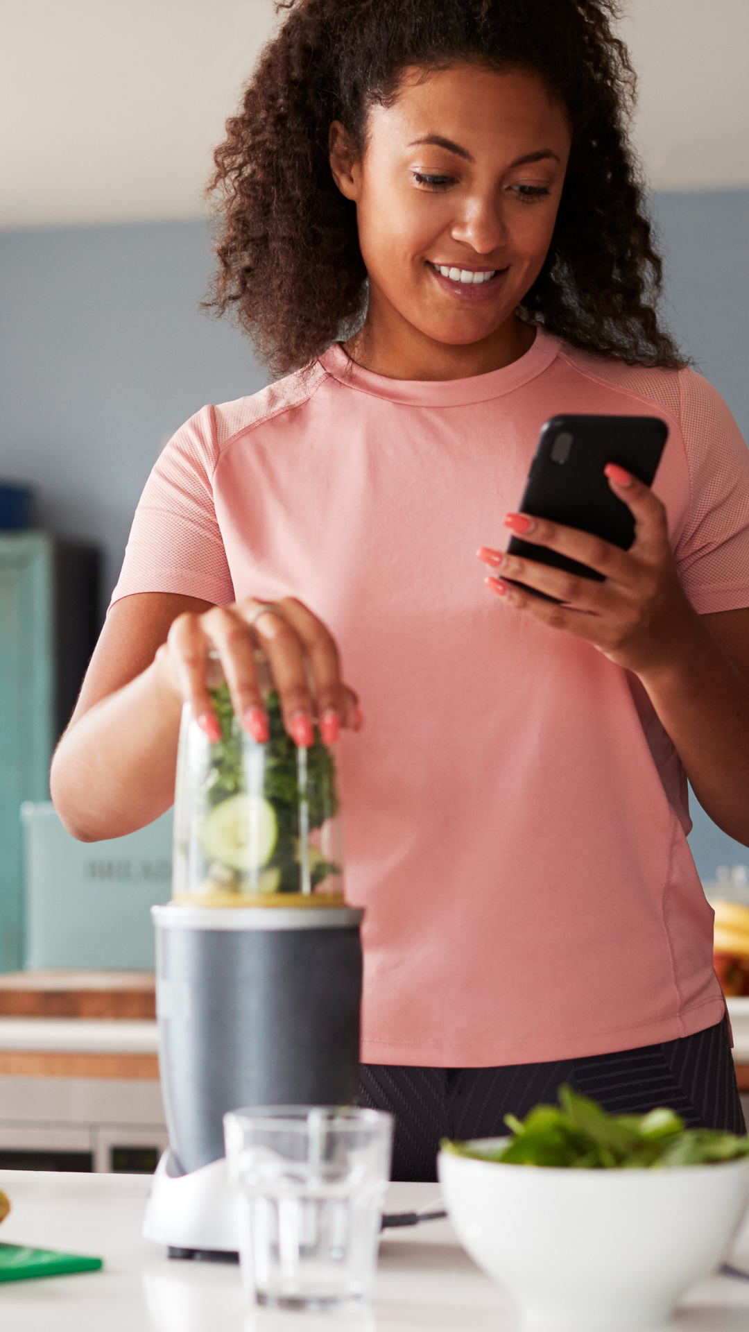 A woman in a pink athletic shirt smiling while looking at her phone, with her hand on a blender container filled with vegetables and a cucumber on the countertop in a kitchen.
