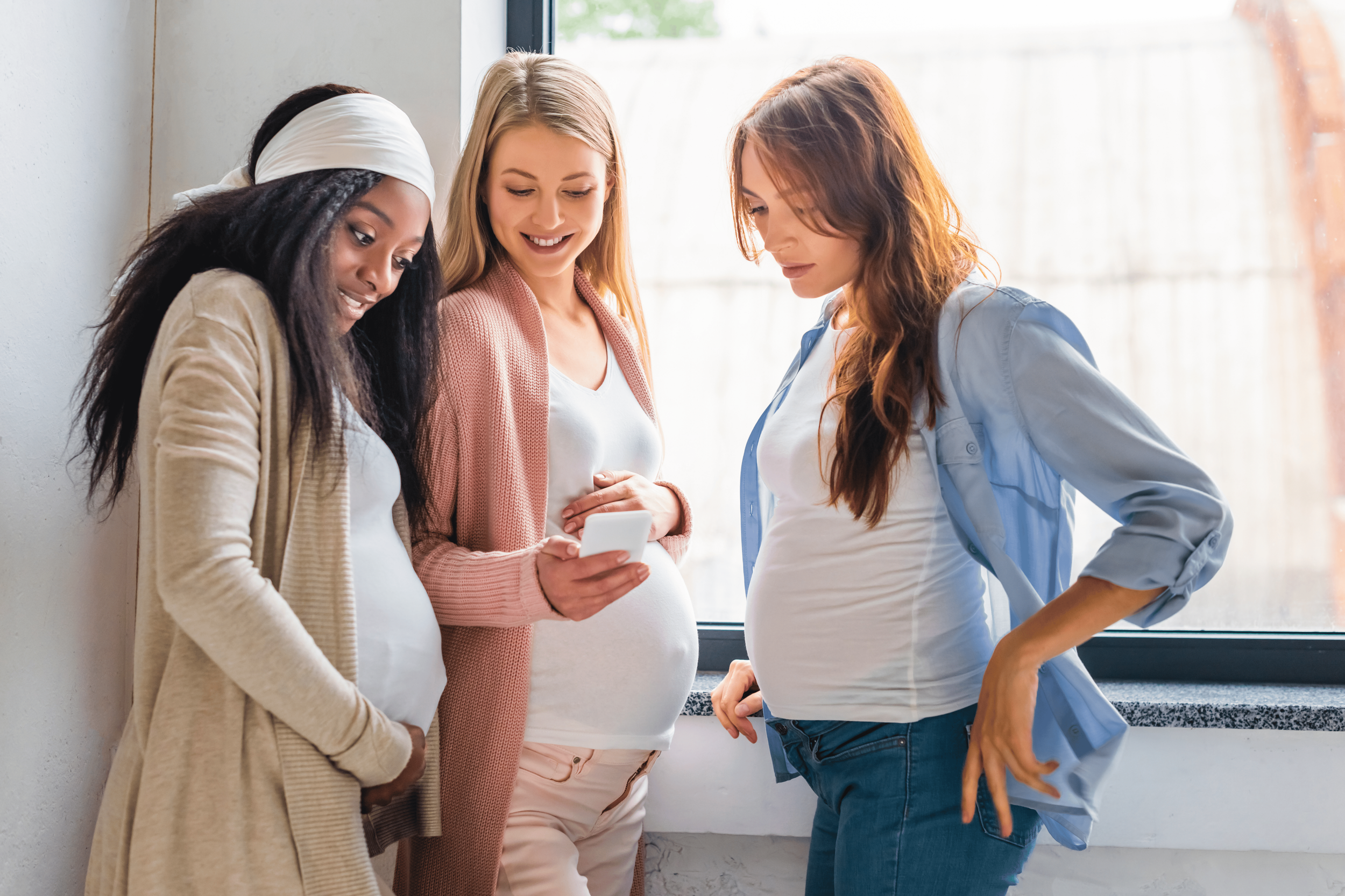 Three pregnant women standing together near a window, looking at a smartphone, smiling, and engaged in conversation.