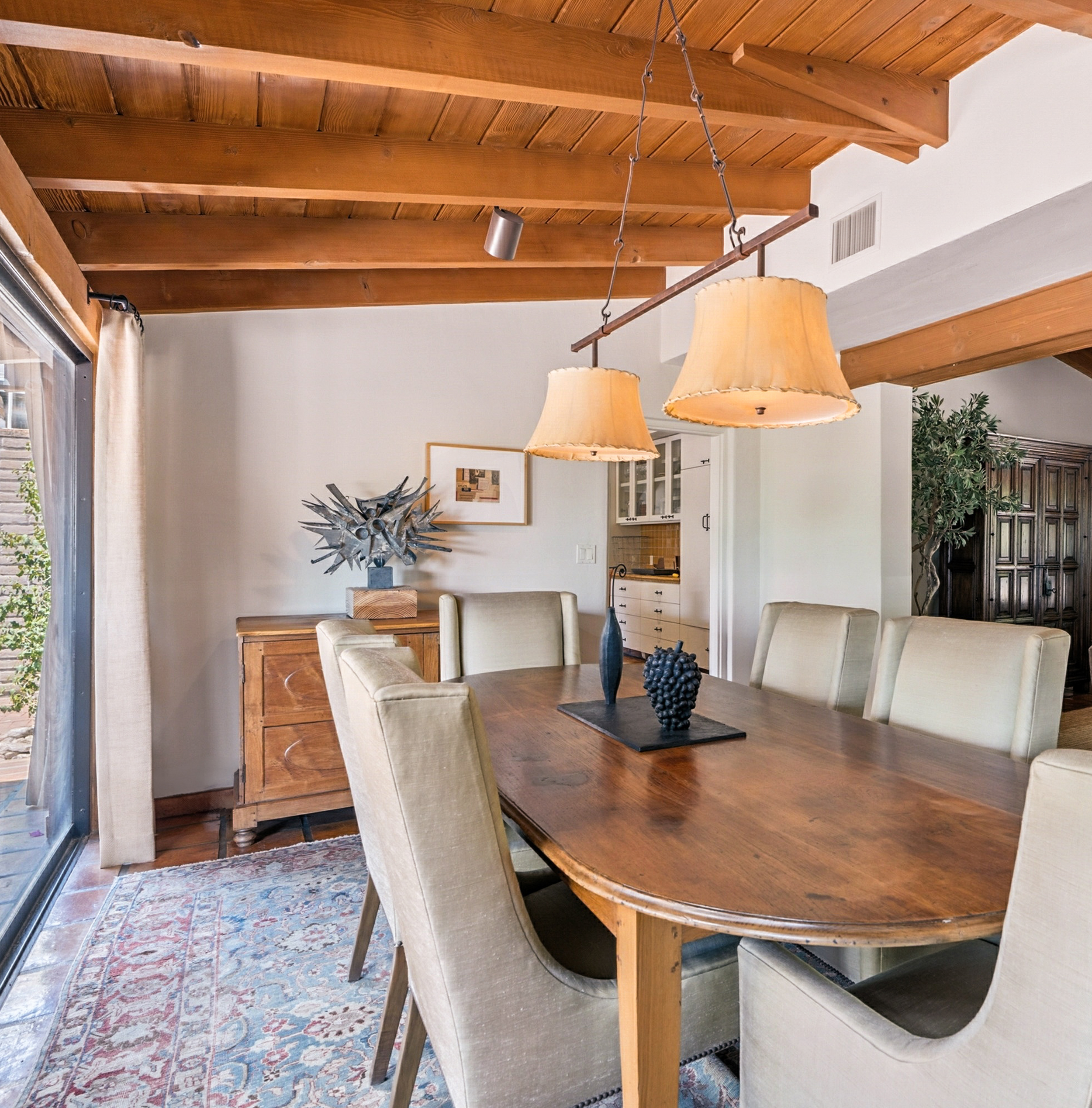 A dining room with a wooden table, six upholstered chairs, a wooden sideboard, a decorative metal sculpture, and a hanging light fixture with beige shades. There is a sliding glass door with curtains and a patterned rug on the floor.
