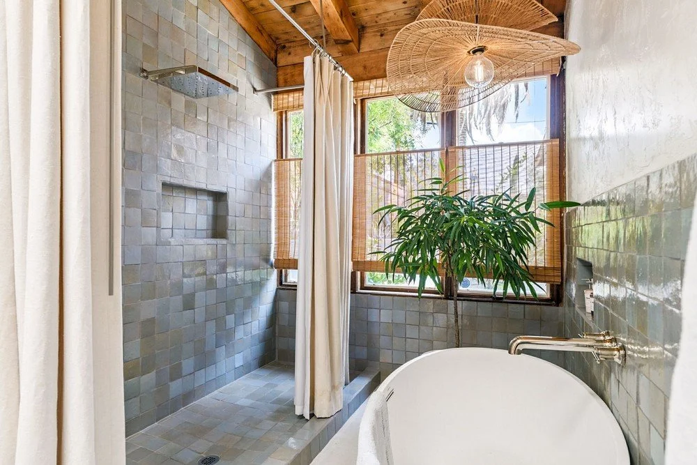 Bathroom with an open shower area, gray tile walls, a bathtub, a large window with bamboo blinds, a potted plant, and a wooden ceiling with woven light fixtures.