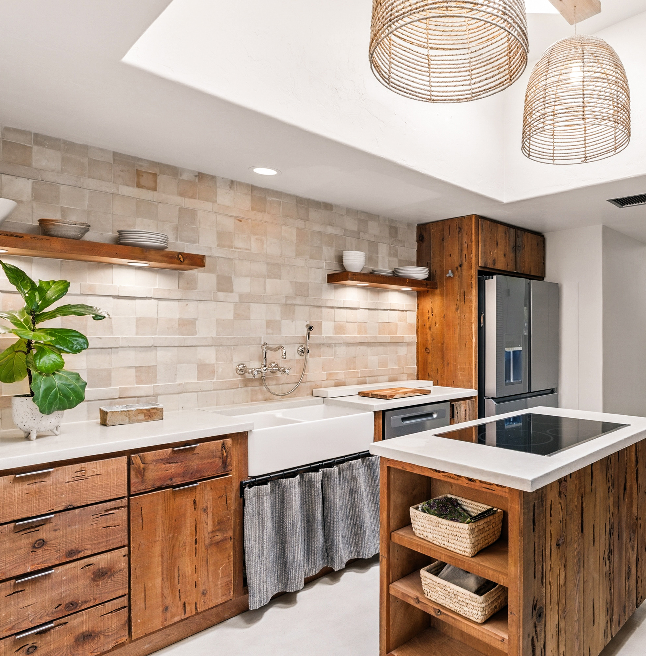 Kitchen with wooden cabinets, white farmhouse sink, gray appliances, open shelves with bowls and plates, green potted plant, textured beige backsplash, island with induction cooktop, hanging woven pendant lights.
