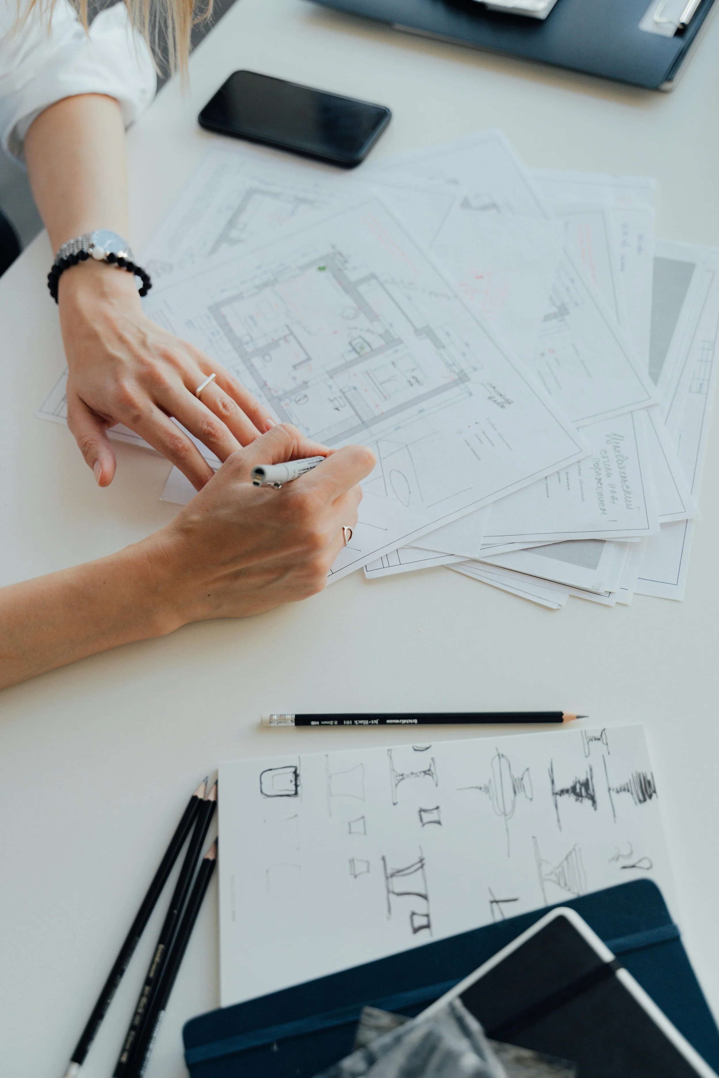 Person working on architectural blueprints at a table with a smartphone, pencils, and sketches of fountain designs.