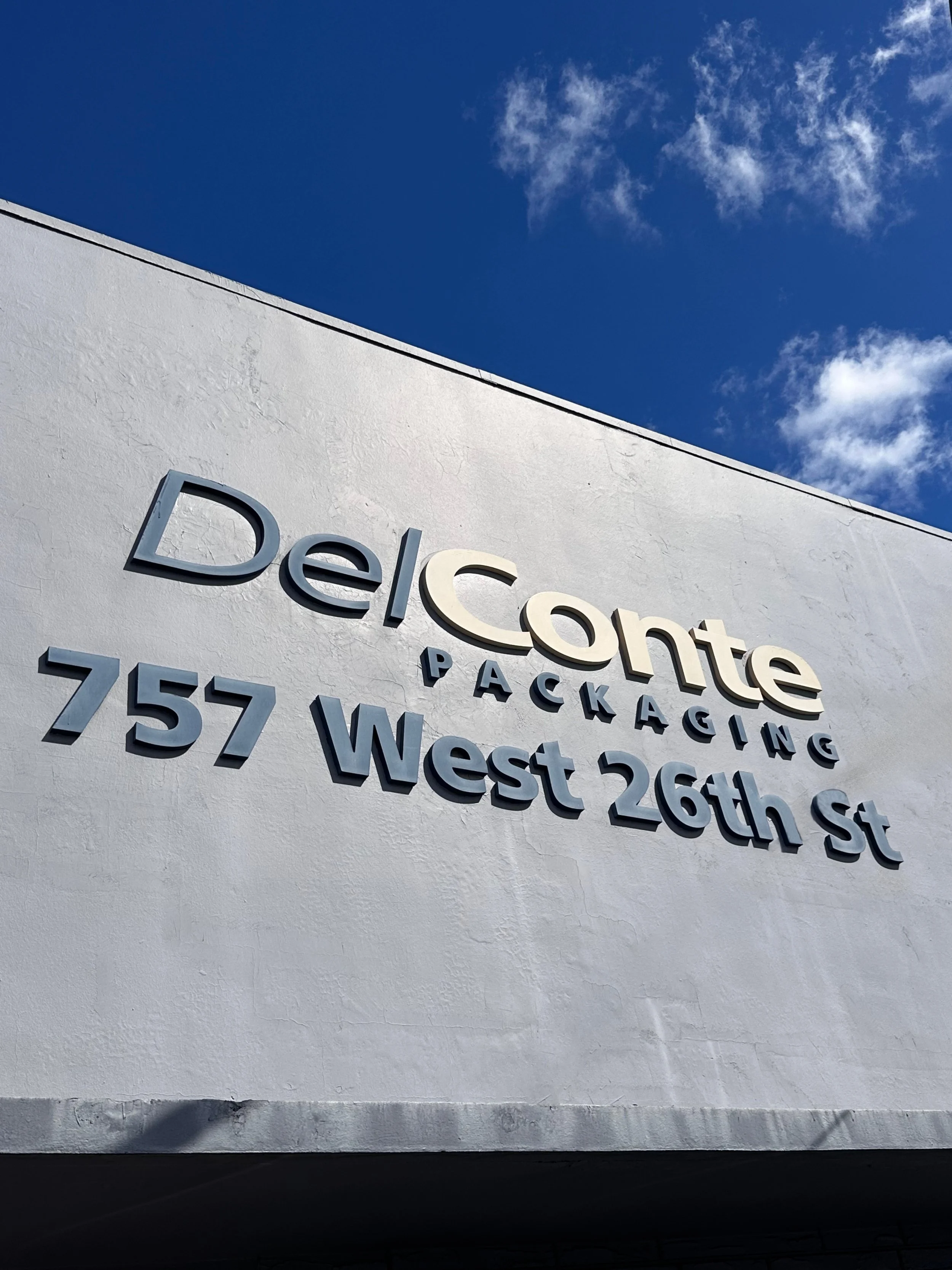 Sign on a white building reading "DelConte Packaging" with an address "757 West 26th St" against a blue sky with some clouds.