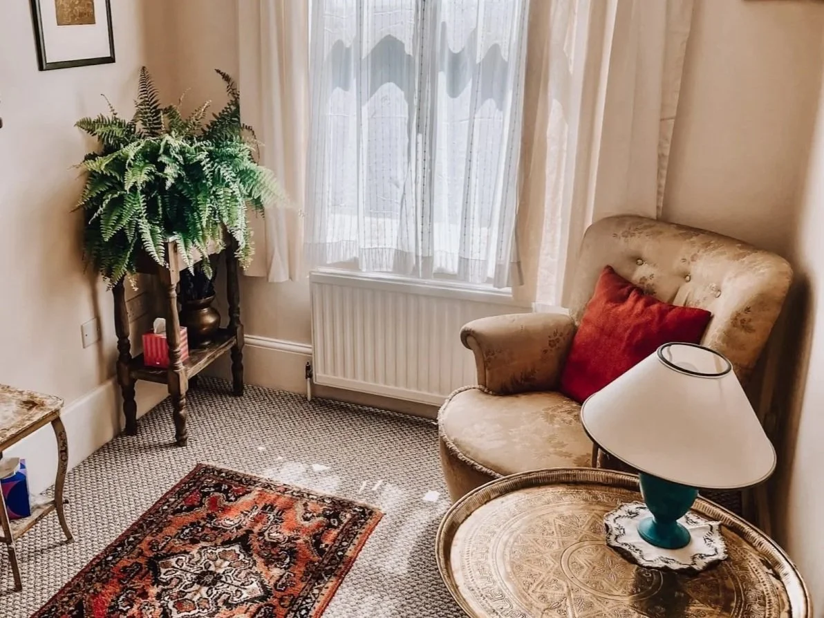 A cozy living room corner with a beige armchair that has a red pillow, a side table with a lamp, a window with sheer curtains, a small table with a potted fern plant, and a patterned rug on the floor.