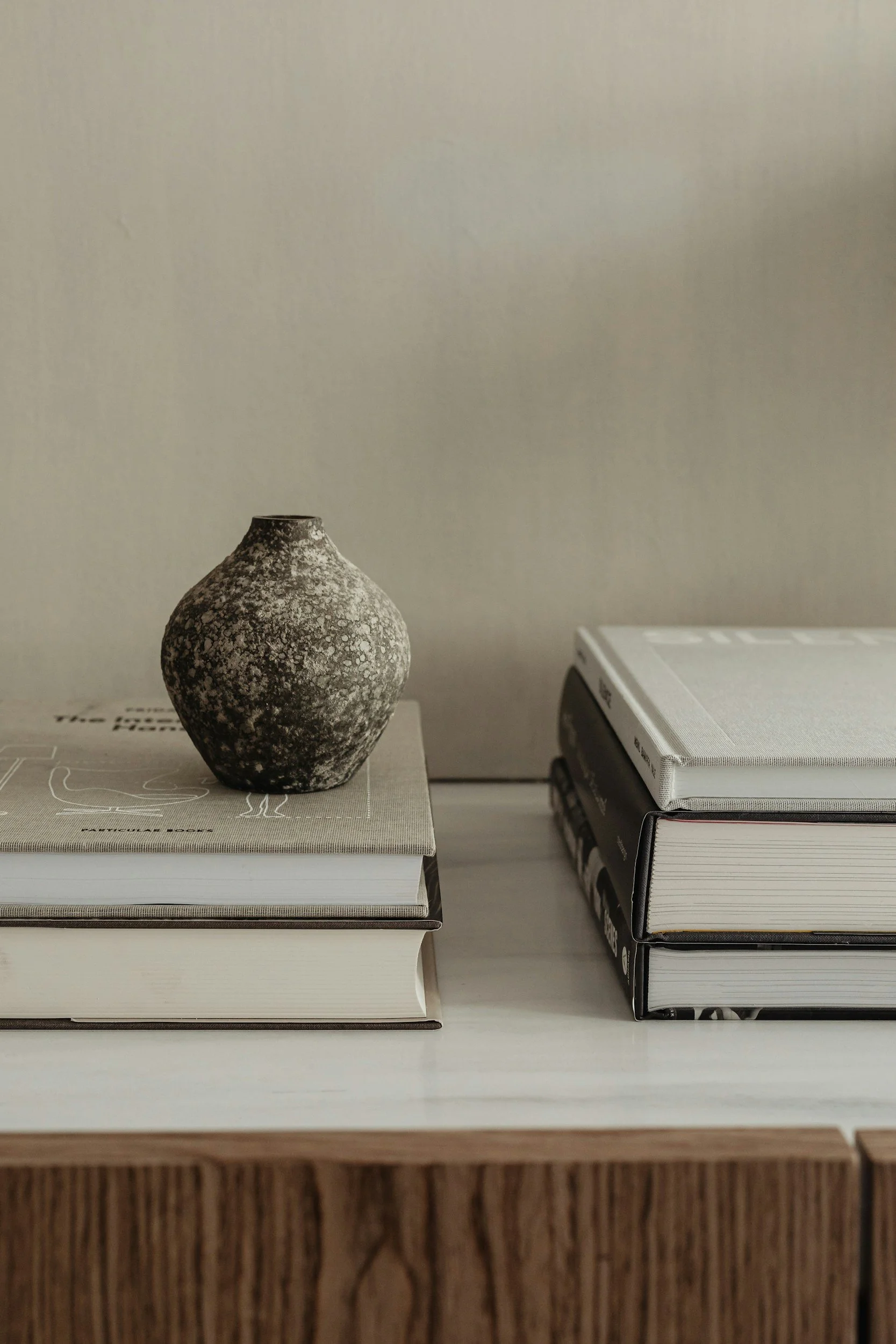 A small, textured black and white ceramic vase is placed on top of two stacked books on a table. To the right, there are three more stacked books with gray covers. The background is a plain, light-colored wall.