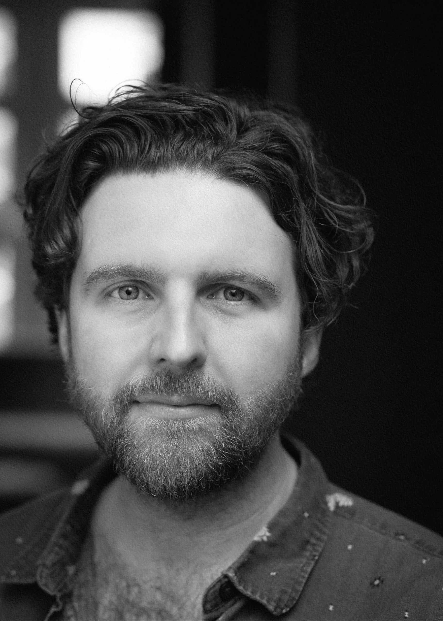 Close-up black-and-white portrait of a man with curly hair, a beard, and light-colored eyes, looking directly at the camera.