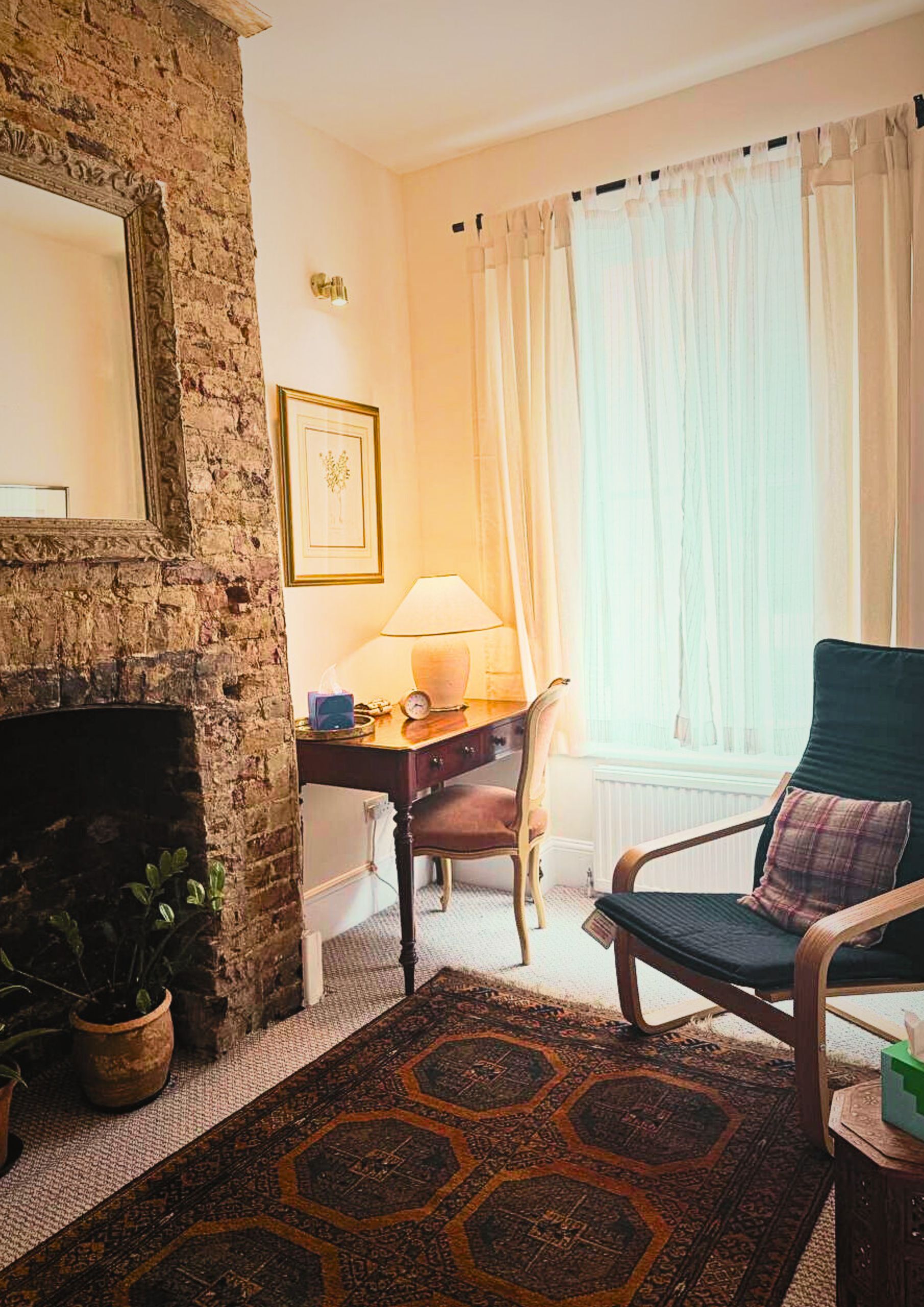 Cozy living room corner with a brick fireplace, a wooden desk with a lamp, a purple upholstered chair, a black armchair with a pillow, a potted plant, a patterned rug, and cream curtains letting in natural light.