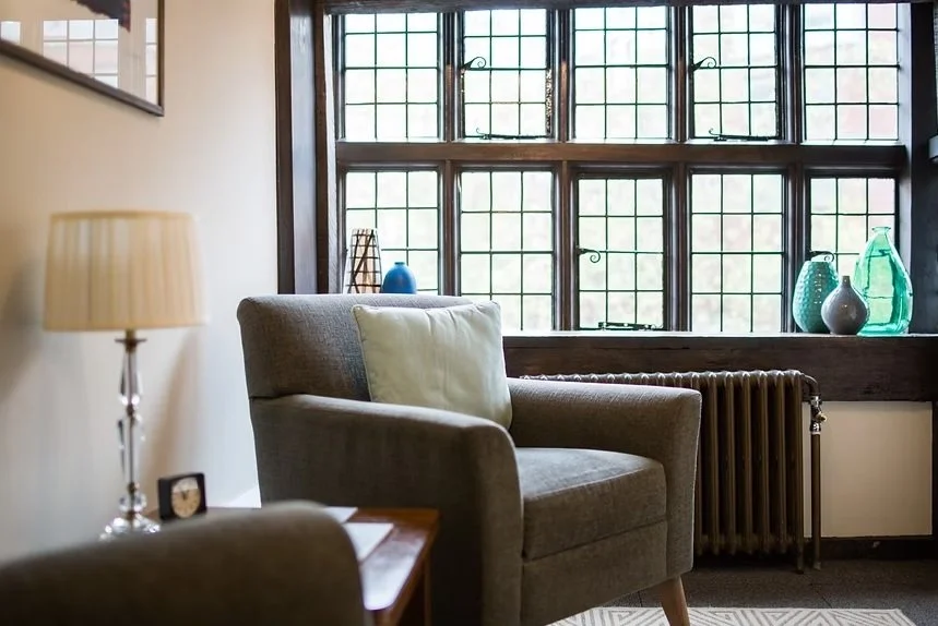 A cozy living room corner with a gray armchair and a cream pillow, a lamp on a small wooden table, large multi-pane windows with decorative glass, and colorful glass vases on the windowsill.