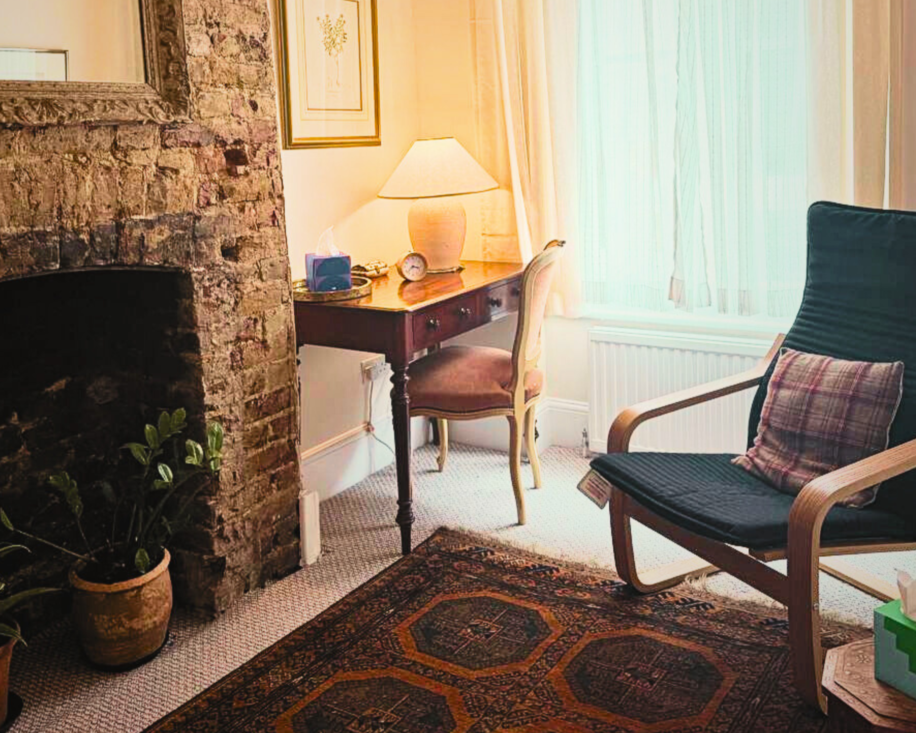 Corner of a cozy room with a brick fireplace, a potted plant, a wooden desk with a lamp, a vintage-style chair, and an armchair with a plaid pillow. Light filters through a window with sheer curtains.