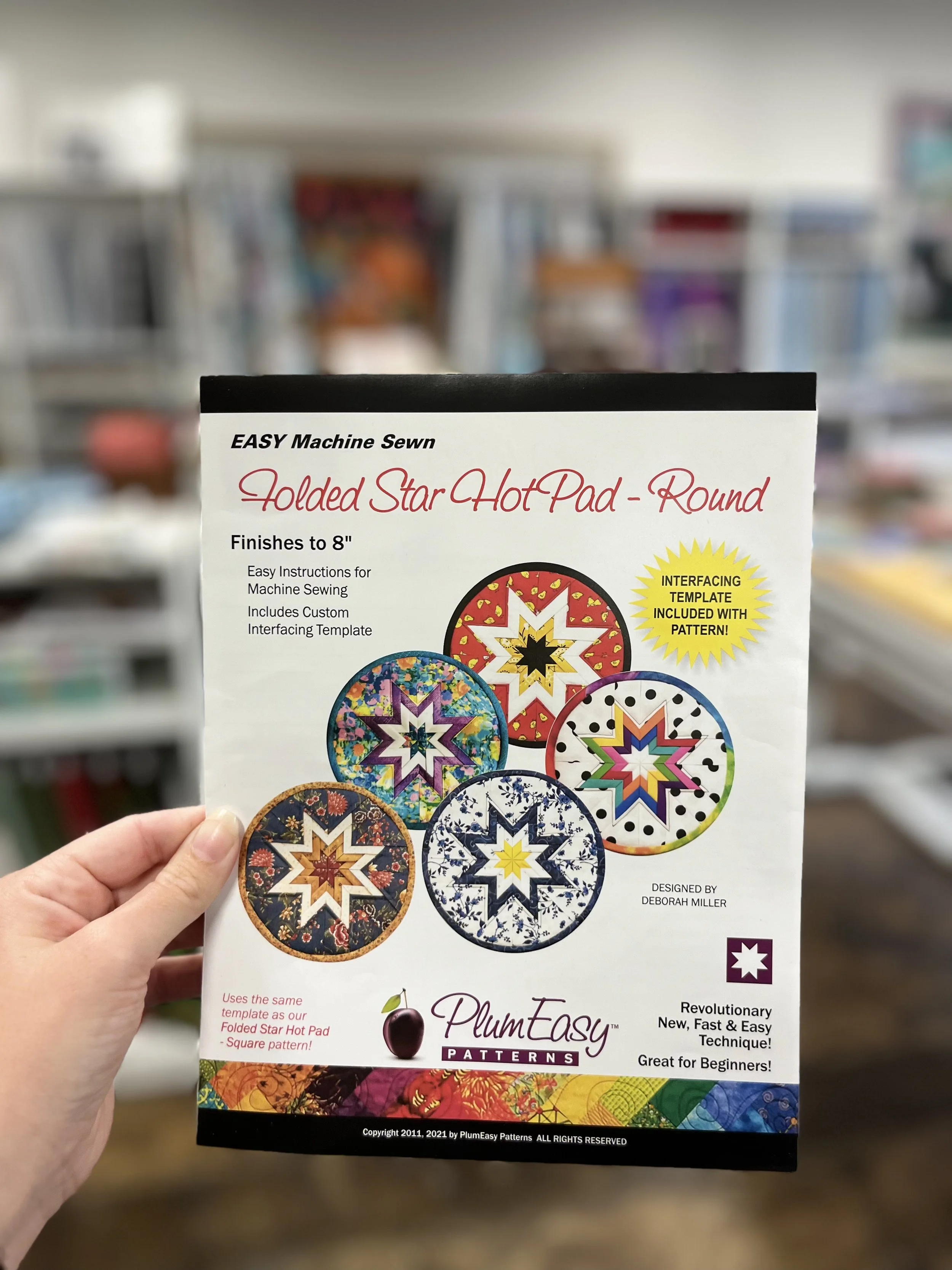 A pack of PlumEasy Patterns Folded Star Hot Pad - Round, showing colorful star patterns on hot pads and text describing ease of machine sewing, pattern details, and designer Deborah Miller.