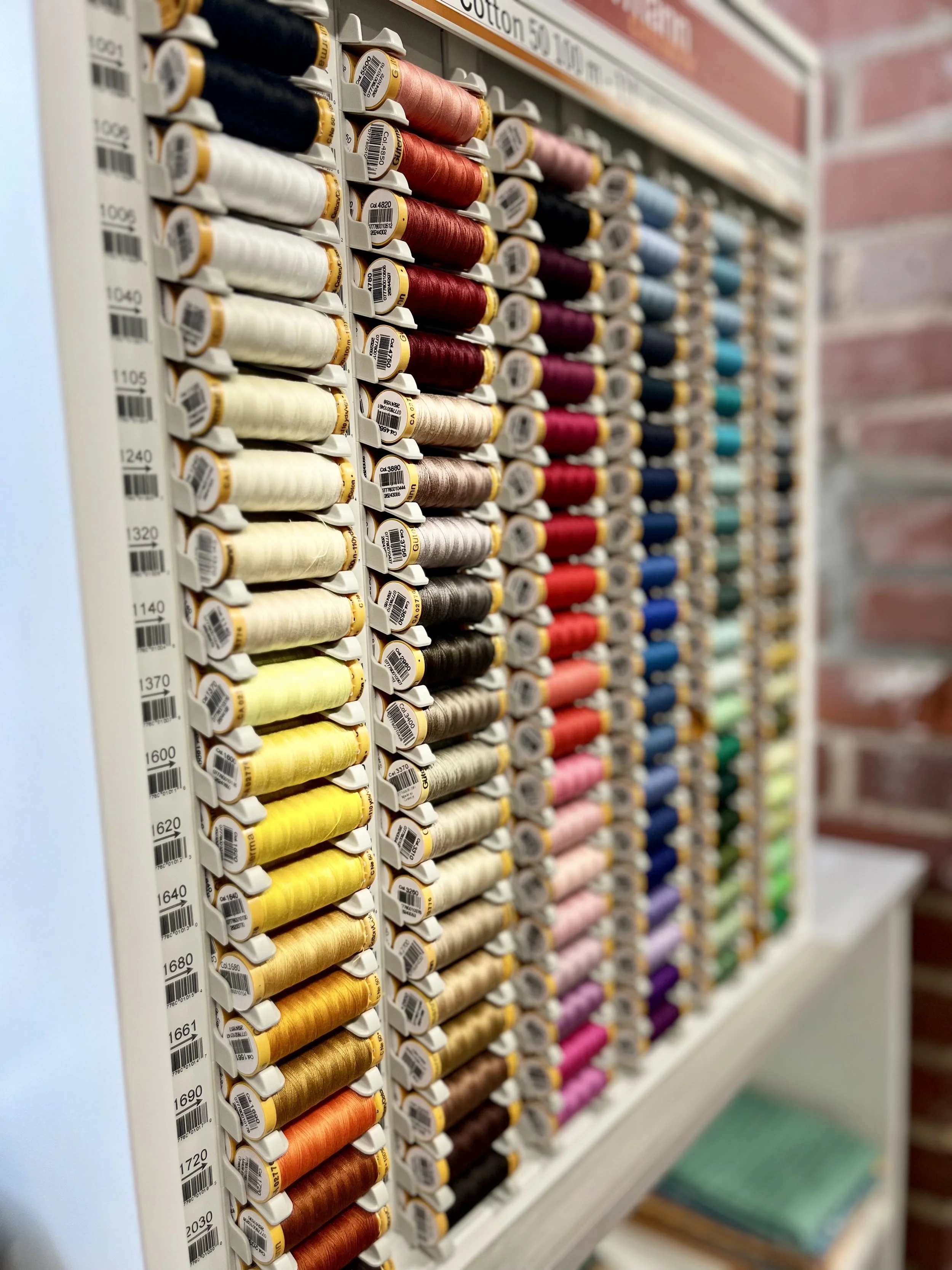 Close-up of a wall-mounted display of multiple rows of colorful sewing machine threads in a craft or fabric store.
