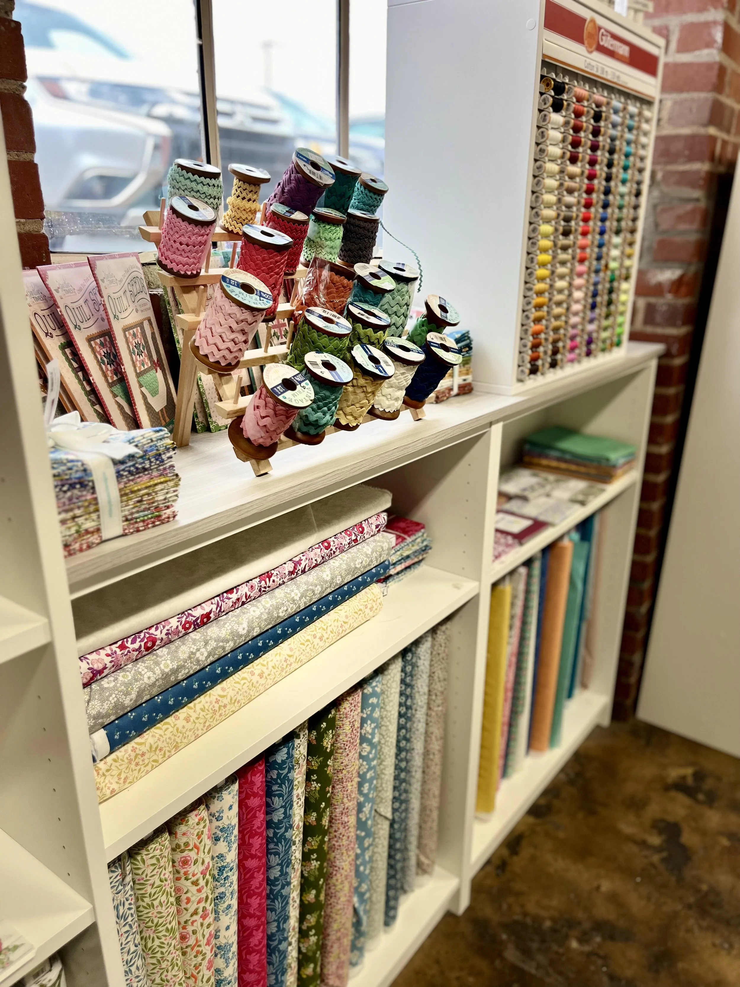 Display of colorful sewing thread spools, quilting patterns, fabric bolts, and a thread color sample chart in a craft store or sewing shop.