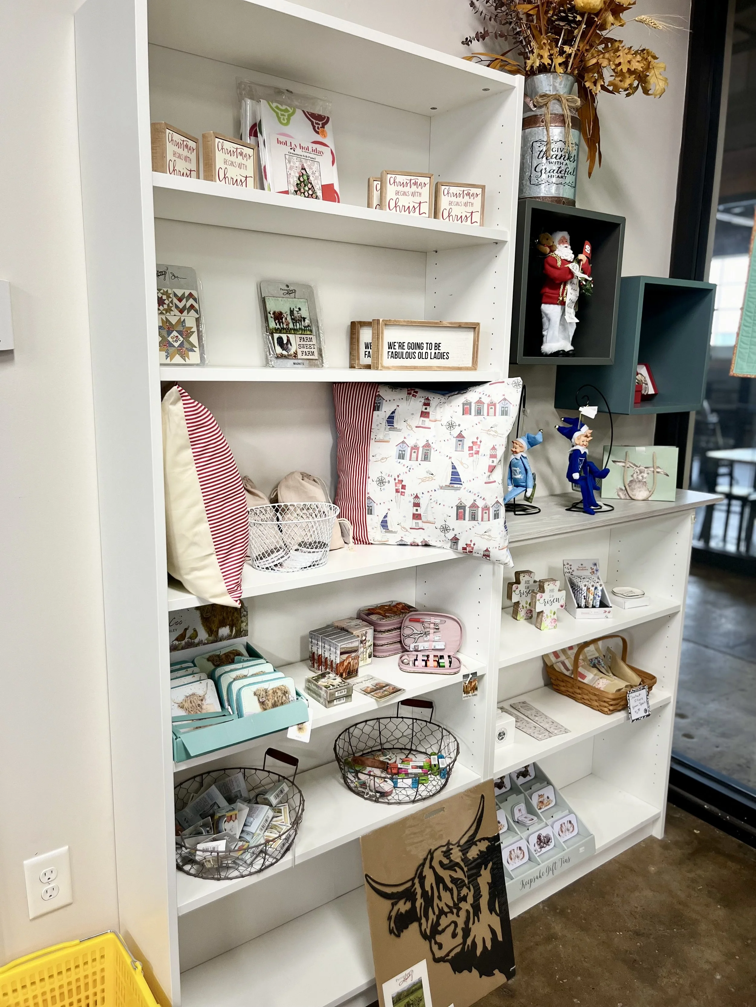 A white bookshelf decorated with Christmas-themed items, pillows, figurines, and small gifts, with a vase of dried flowers on top, near a window.