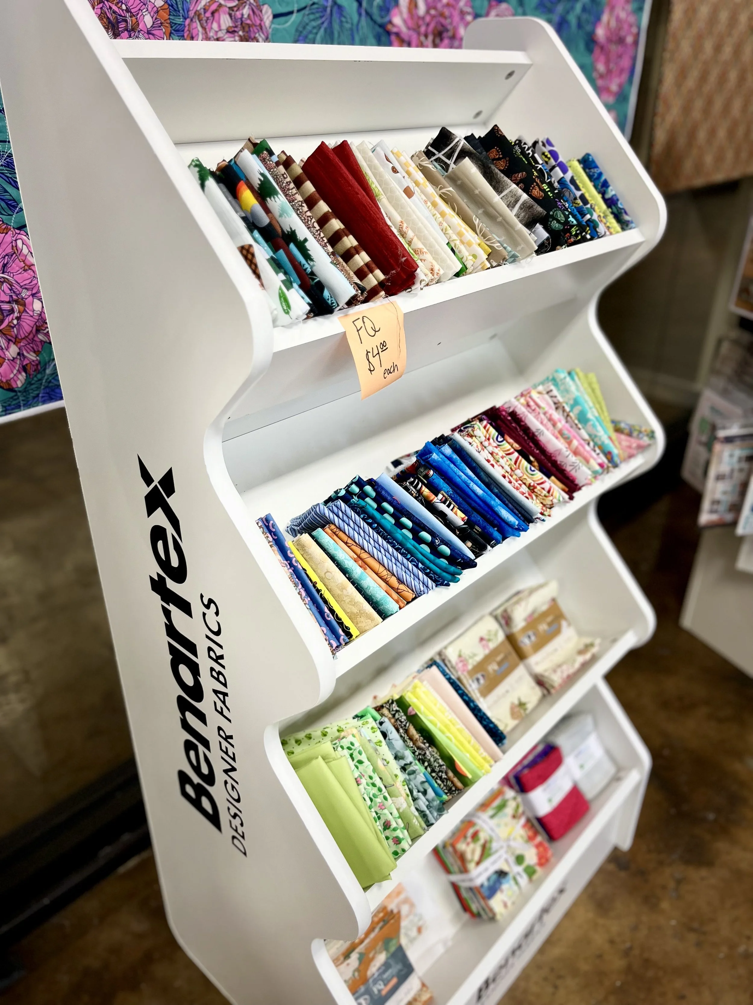 A white display rack holding various folded fabrics in multiple colors and patterns, with a sign indicating each piece costs $4.00.