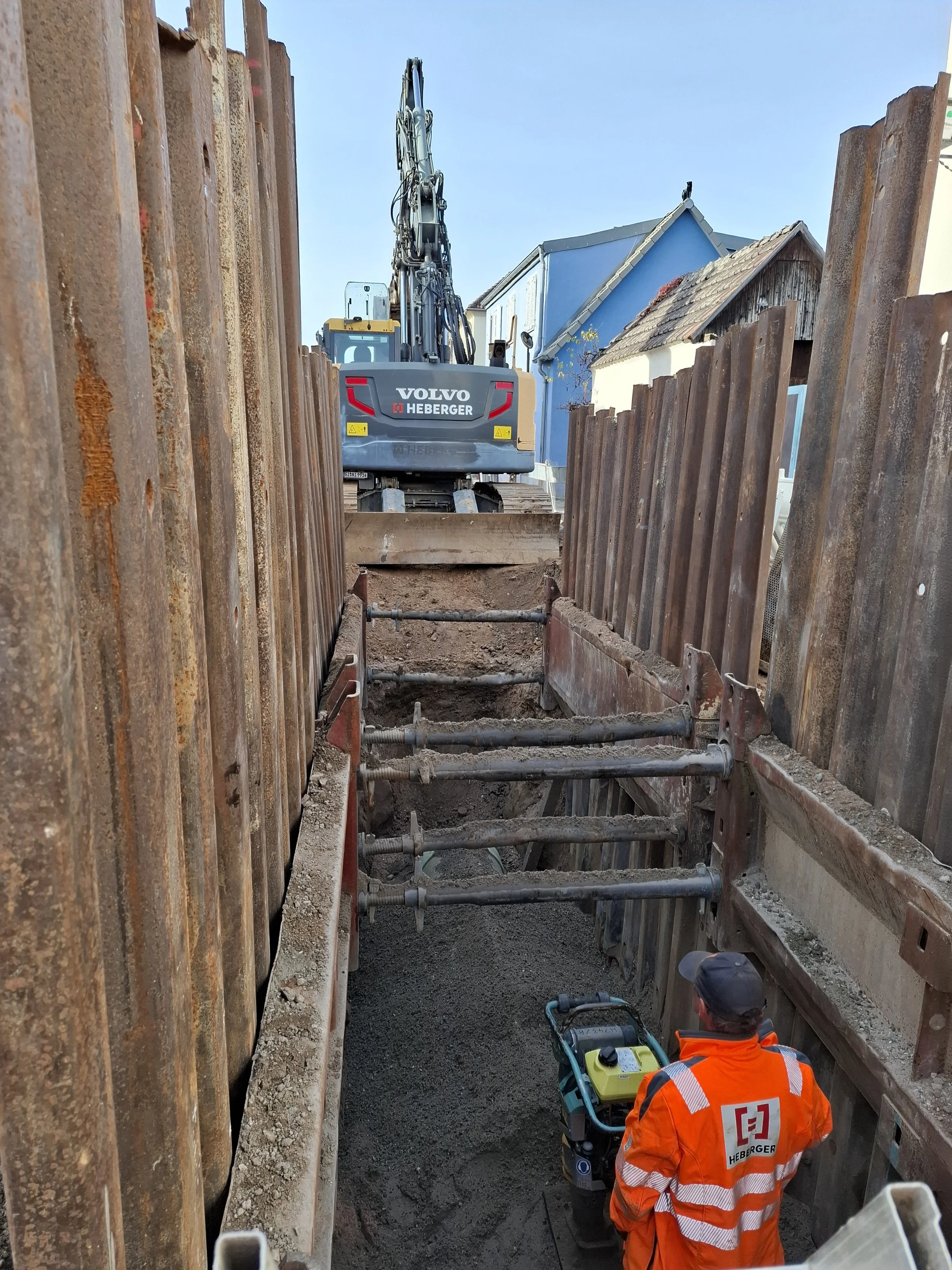 Bauarbeiter beim Verlegen von Rohrleitungen in einem tiefen Graben, umgeben von Baustahlwänden, mit einem Bagger im Hintergrund.