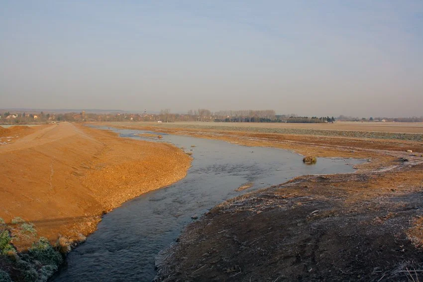 Ein Fluss, der durch ein ländliches Gebiet fließt, umgeben von Feldern und einer kleinen Siedlung im Hintergrund bei Sonnenauf- oder untergang.