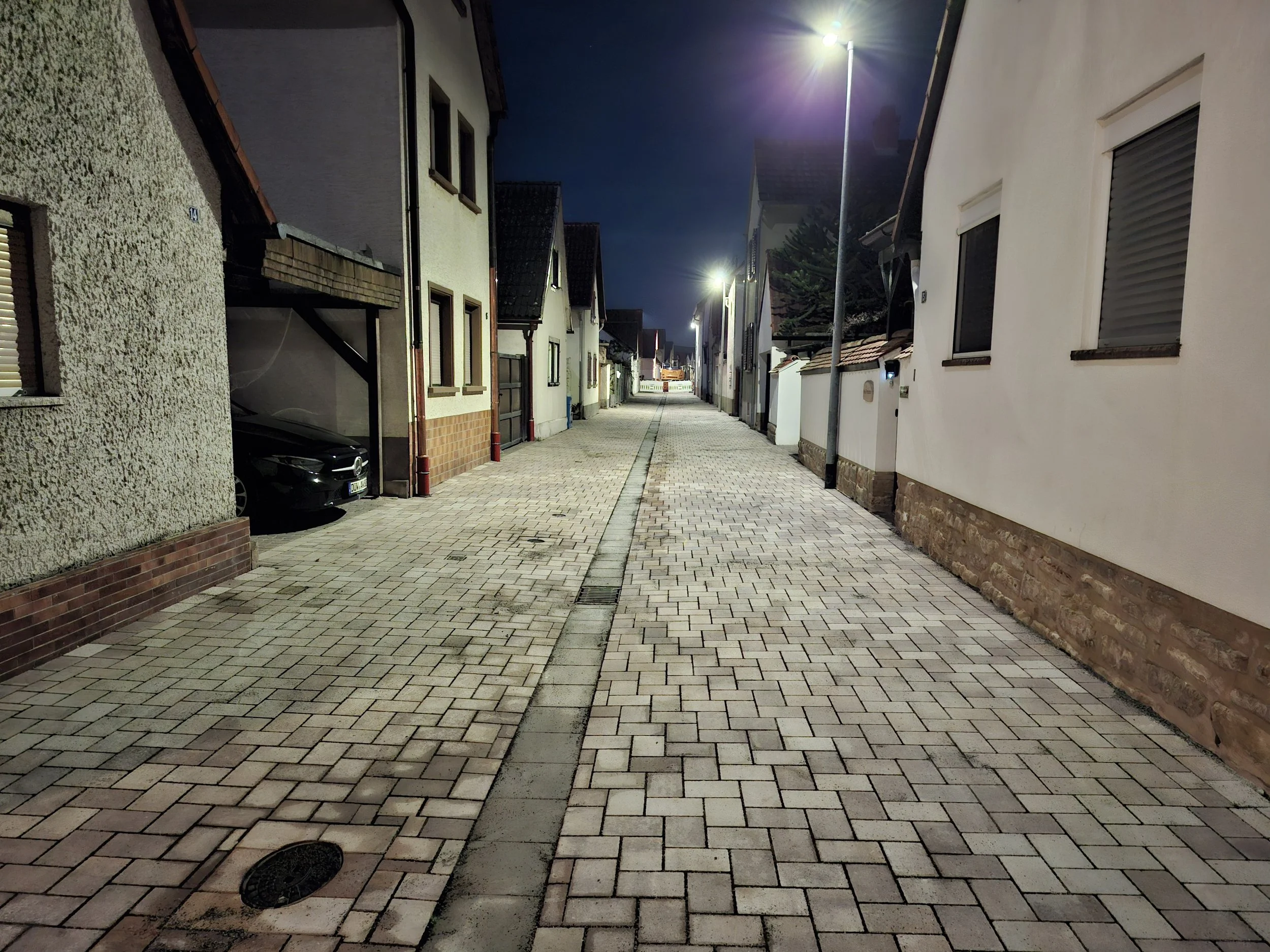 Leere nächtliche Fußgängerzone mit gepflastertem Gehweg, Häuser auf beiden Seiten und Straßenlaternen, die den Weg beleuchten.
