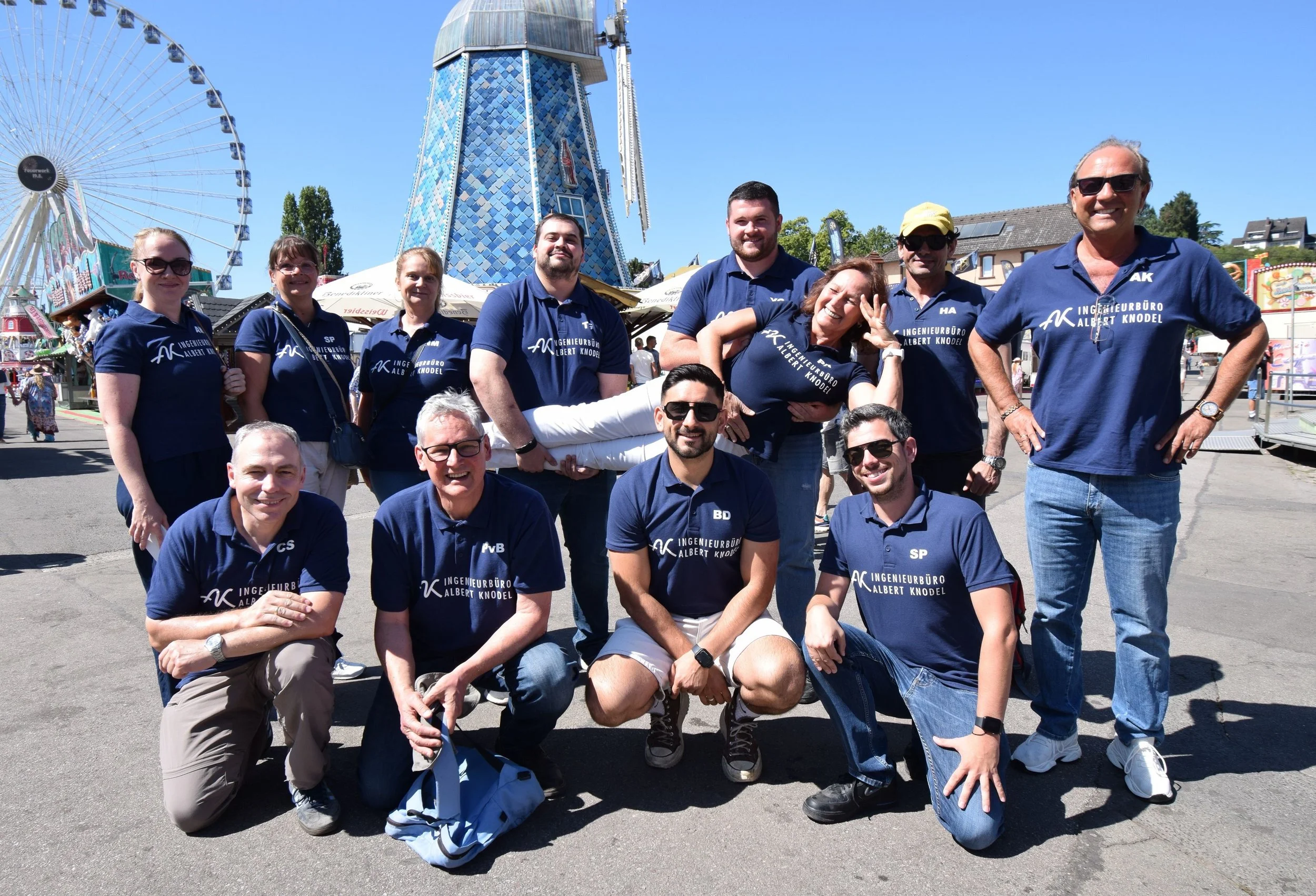 Gruppenumarmung von zehn Personen auf einem Volksfest, einige lachen, andere posieren. Im Hintergrund fahrendes Riesenrad und ein blaues Windrad. Die Gruppe trägt dunkelblaue T-Shirts mit weißen Schriftzug, einige tragen Sonnenbrillen, die meisten sind Männer, ein Mädchen ist lächelnd, eine Person wird hochgehalten.