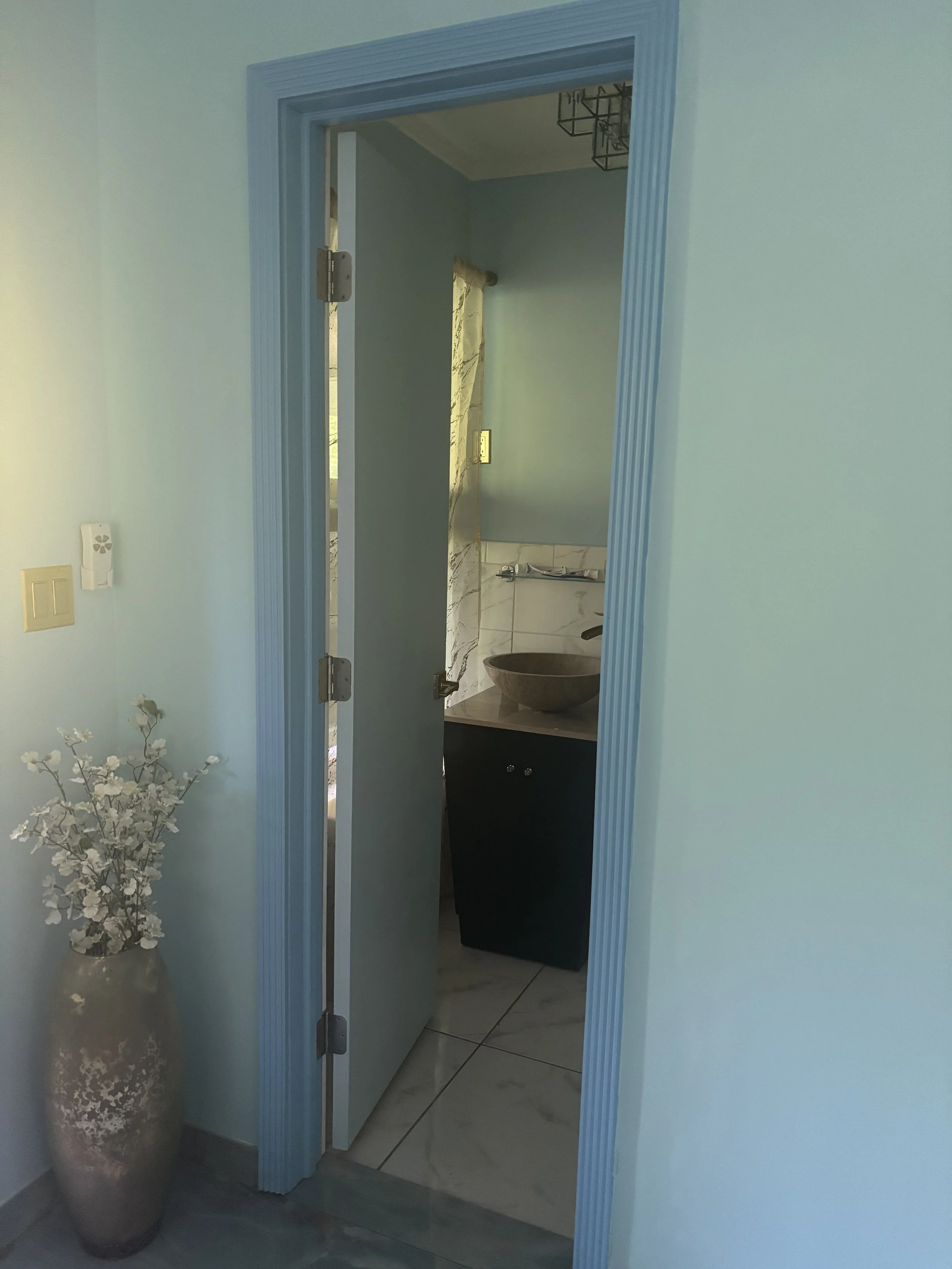 View into a small bathroom through a slightly open pastel blue door, with a vessel sink on a dark cabinet, marble walls, and tile flooring.