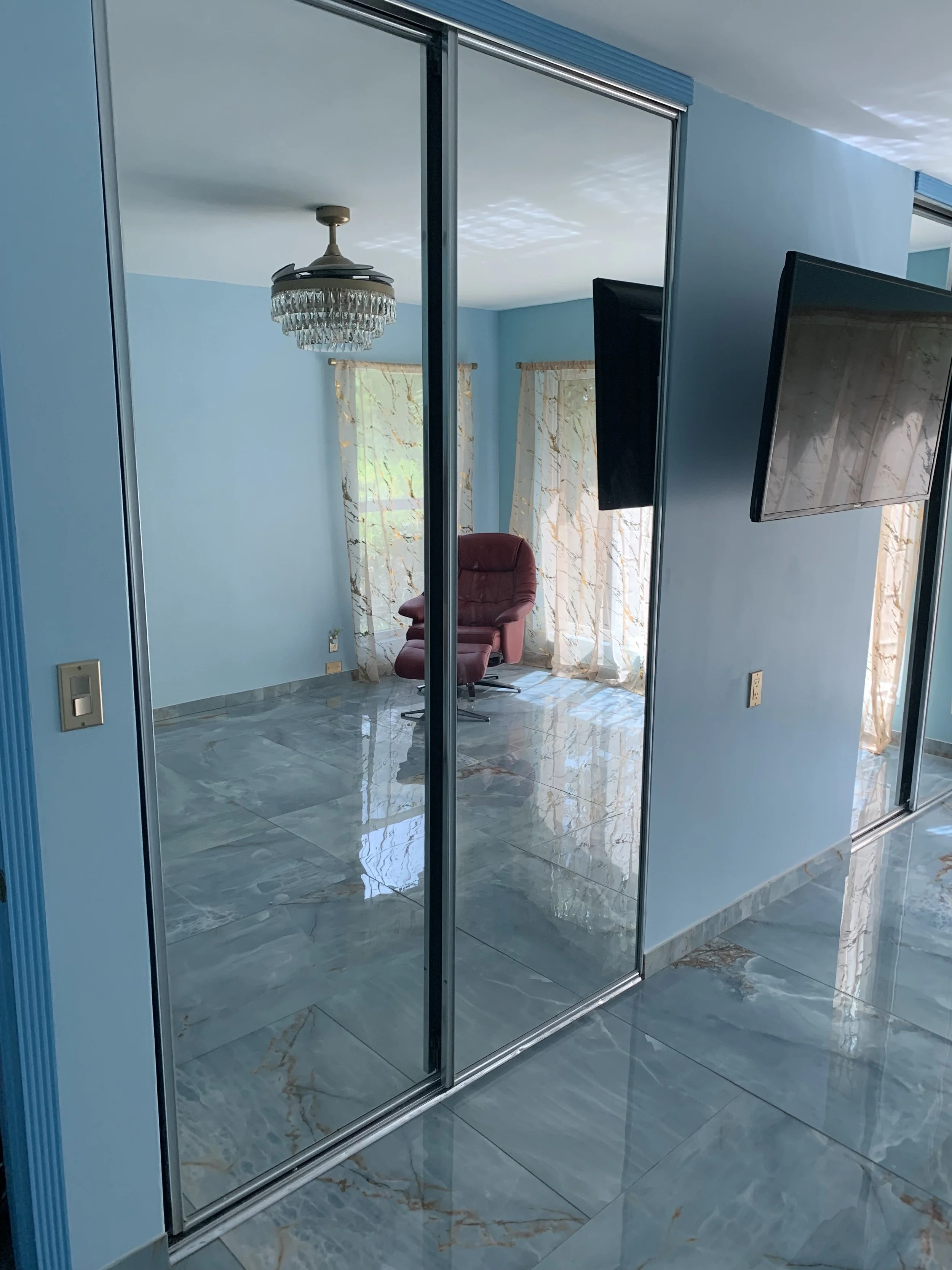 Reflection of a blue-walled room with a burgundy recliner chair, cream curtains with leaf pattern, and a chandelier in the mirrored closet door.